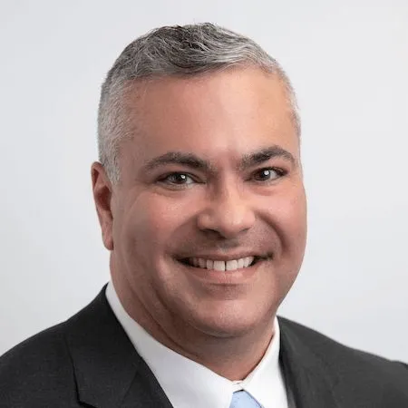 Professional headshot of a smiling man in a black suit and white shirt, gray background