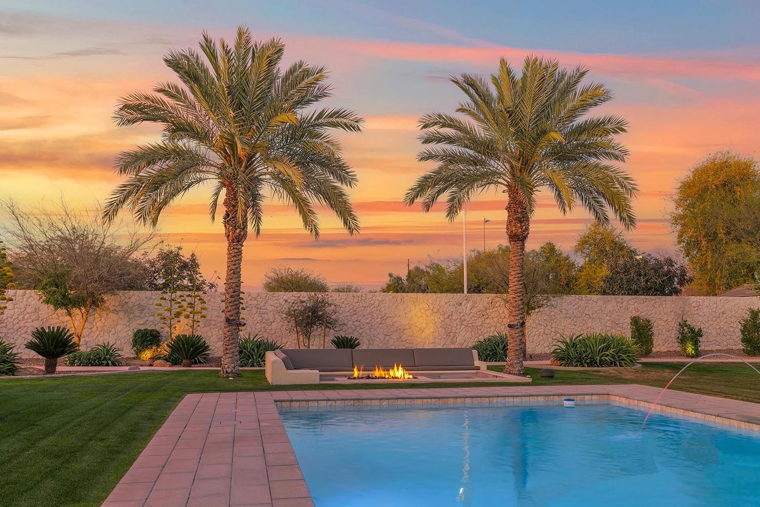 Cozy living room with hardwood floors, large windows overlooking a lake, wood-paneled ceiling, floral sofa, armchairs, wooden coffee table with flowers, and red curtains, in a lakeside cabin or home.
