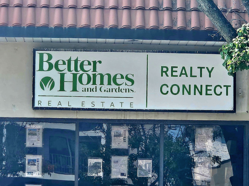Office reception area with a realtyconnect usa sign, cubicle desks, office equipment, and a digital screen displaying webinar announcement.