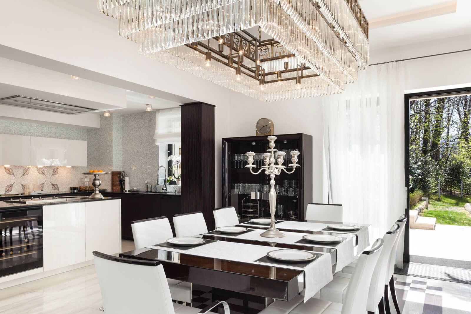 Modern dining room with a black and white color scheme, chandelier, elegant table setting, and view of a lush garden through large glass door.