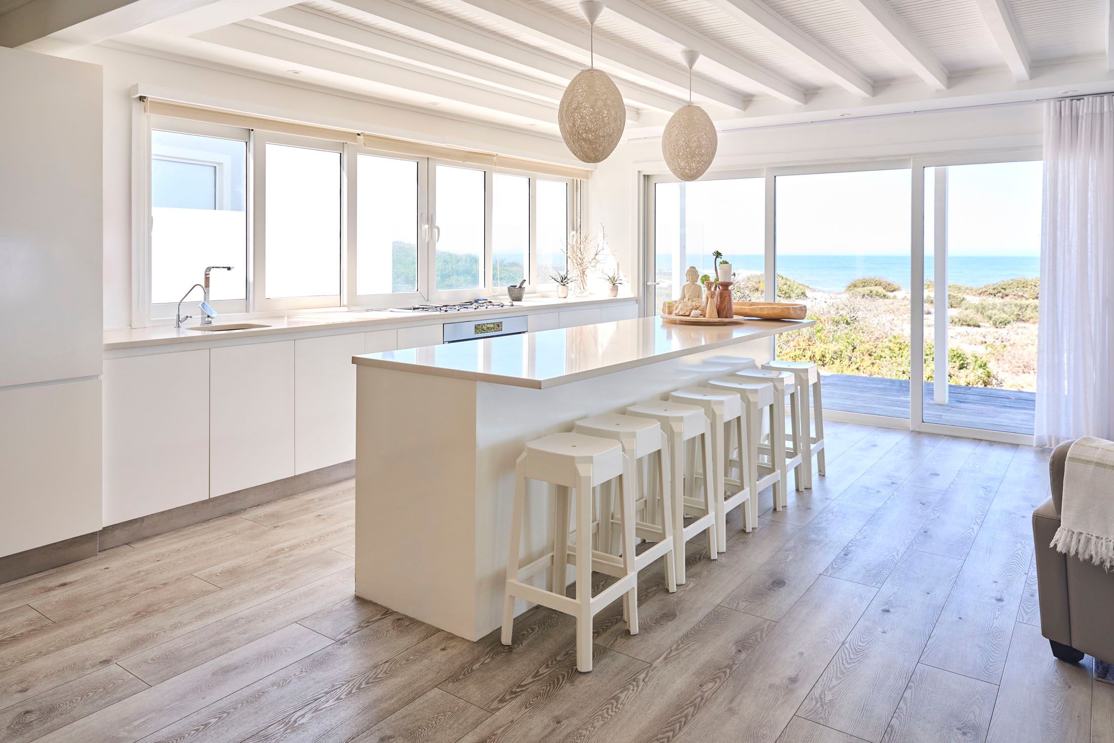 Modern minimalist white kitchen with large ocean view windows, featuring a central island with bar stools, sea-inspired decor, and natural light filtering through sheer curtains.