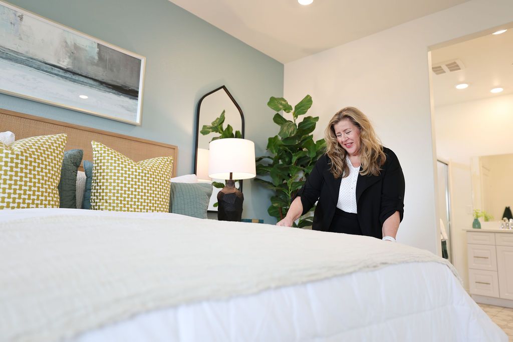 Woman making a bed in a modern, well-decorated bedroom with colorful pillows, a lamp, and a large plant.
