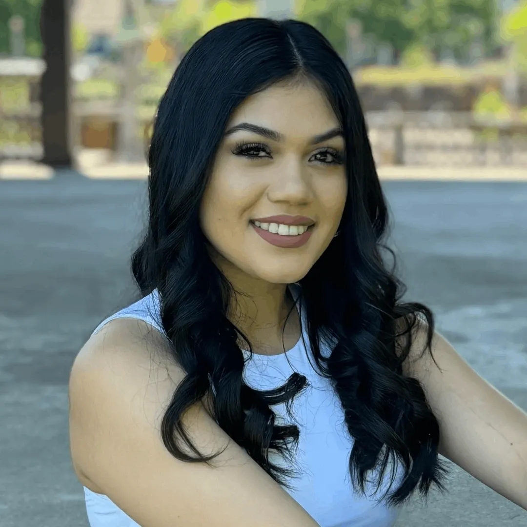 Smiling young woman with long black wavy hair, wearing a white sleeveless top, outdoors in natural light with blurred trees and structures in the background.