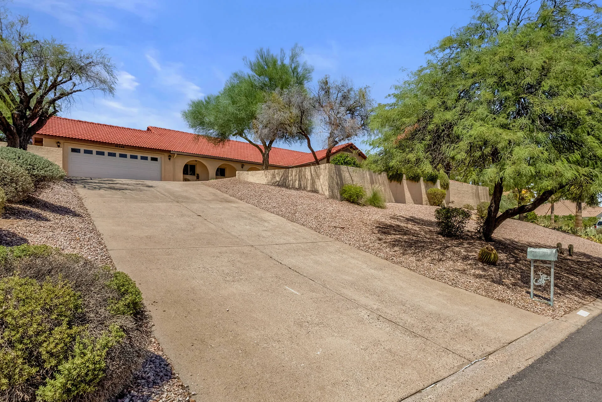 Single-story desert-style house with a tiled roof, surrounded by desert plants including cacti and a large tree, under a bright blue sky.