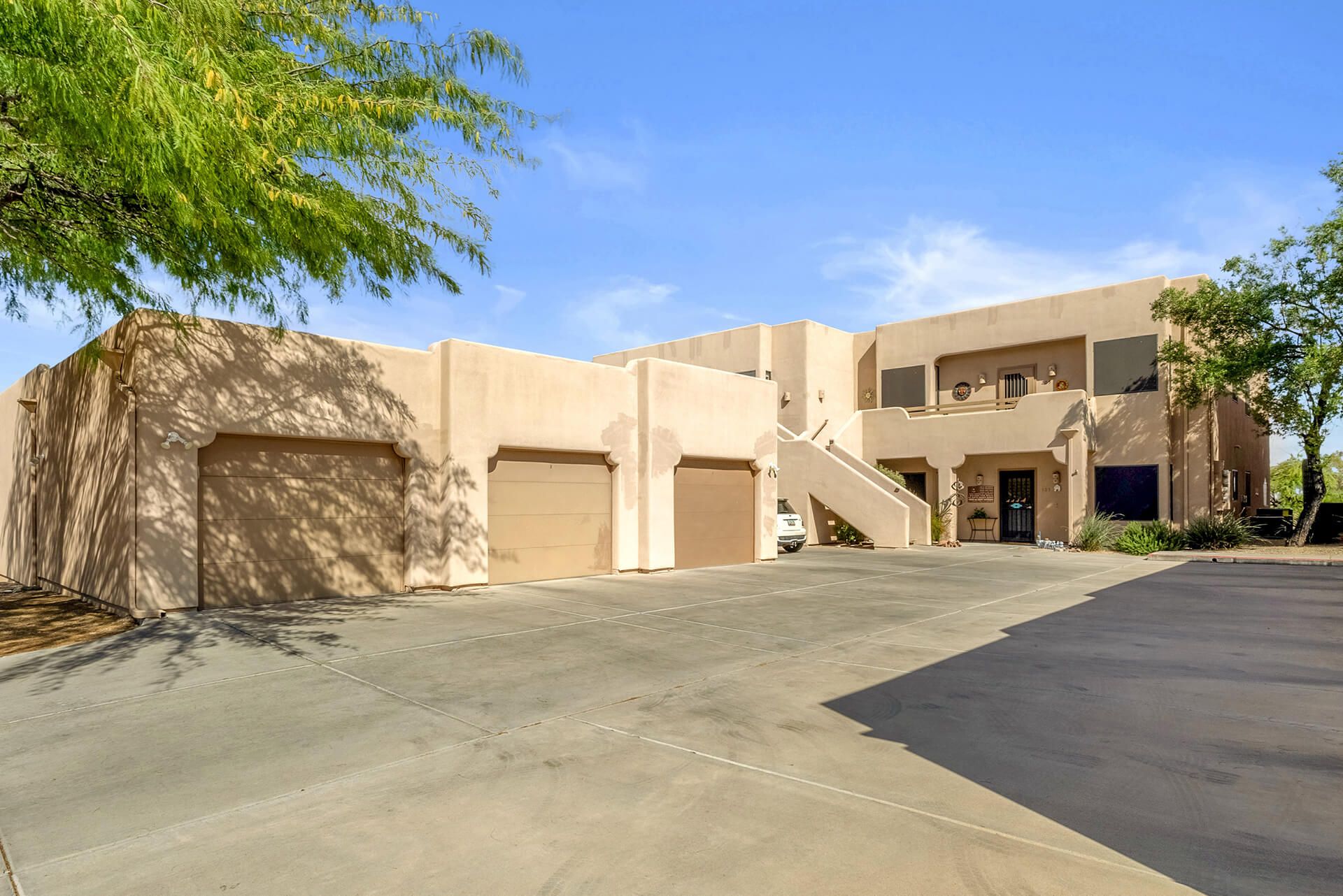 Modern two-story house with beige exterior, attached garage, landscaped front yard, large shade tree, and clear blue sky.