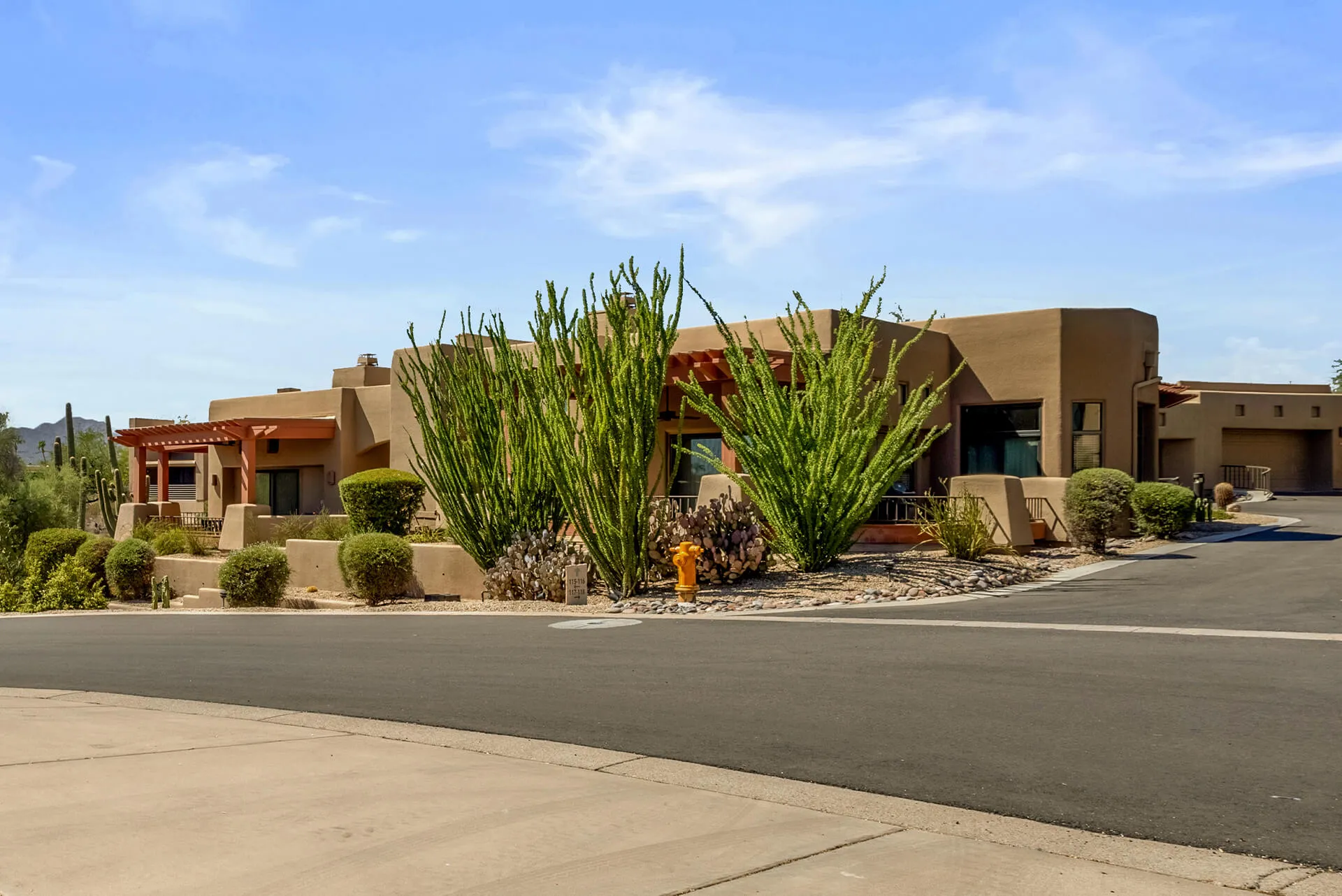 Single-story desert-style house with a tiled roof, surrounded by desert plants including cacti and a large tree, under a bright blue sky.