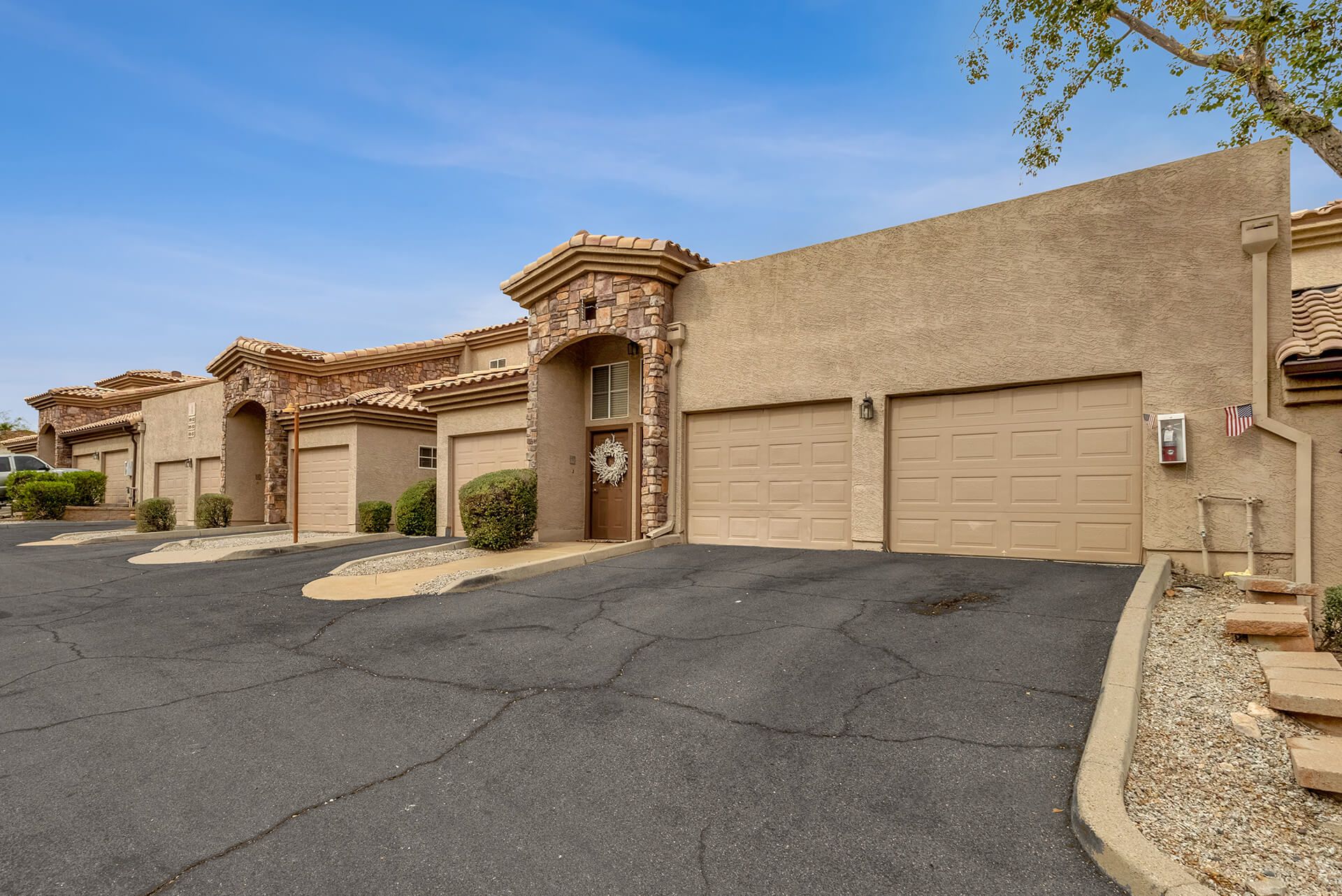 Modern two-story house with beige exterior, attached garage, landscaped front yard, large shade tree, and clear blue sky.