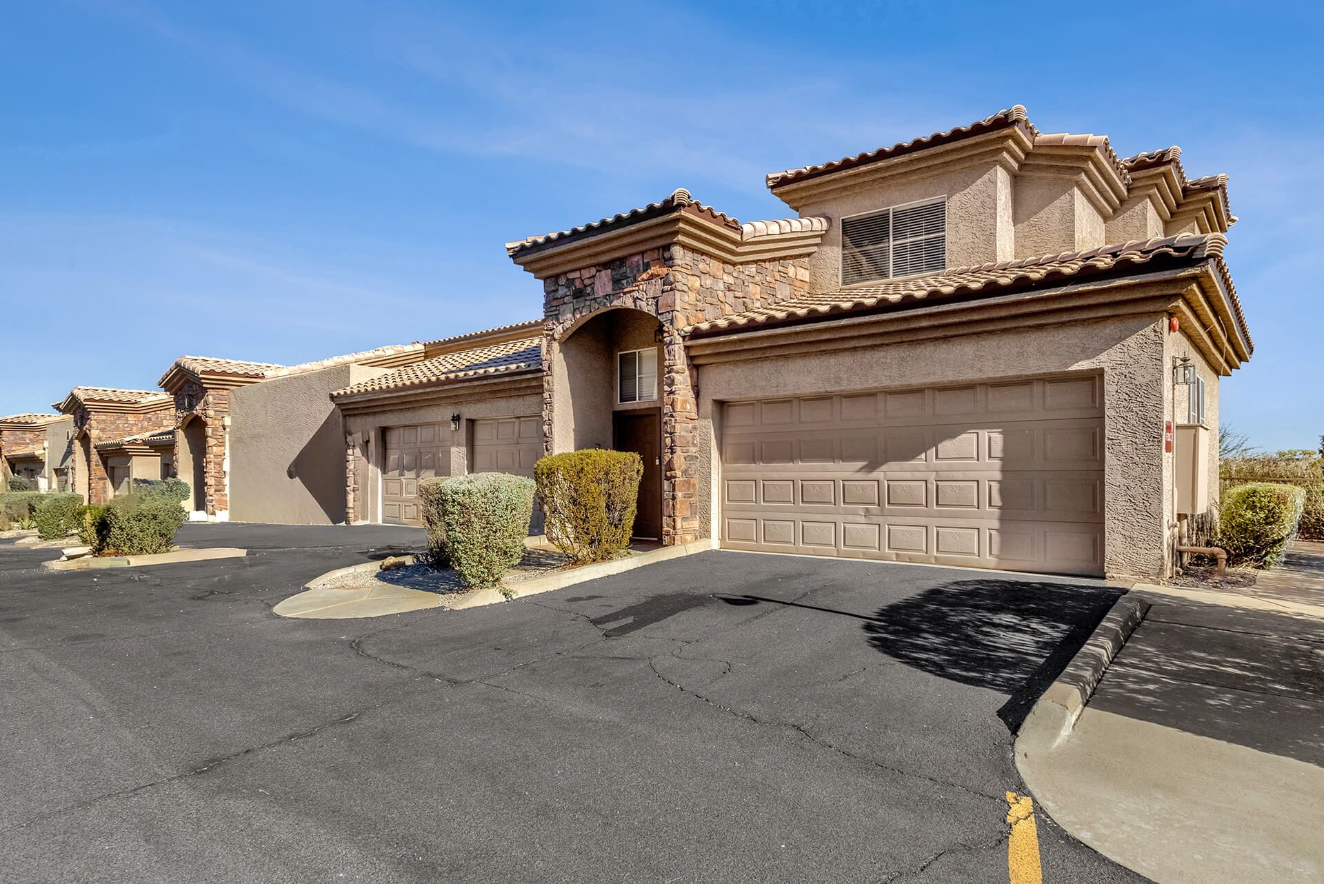 Modern two-story house with beige exterior, attached garage, landscaped front yard, large shade tree, and clear blue sky.