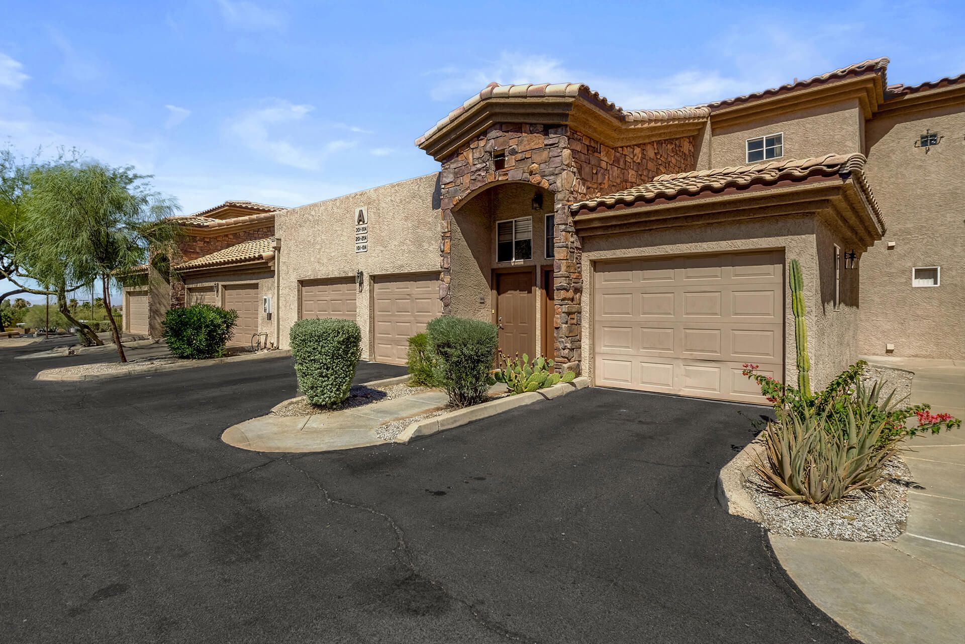 Modern two-story house with beige exterior, attached garage, landscaped front yard, large shade tree, and clear blue sky.