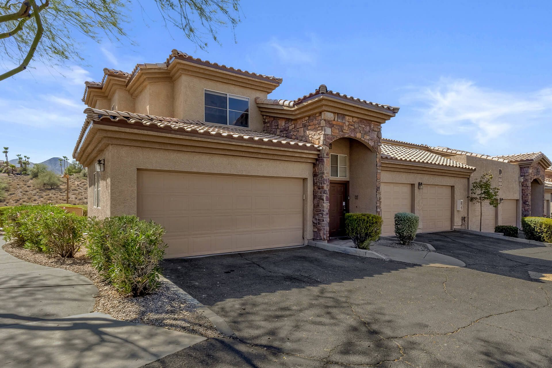Modern two-story house with beige exterior, attached garage, landscaped front yard, large shade tree, and clear blue sky.