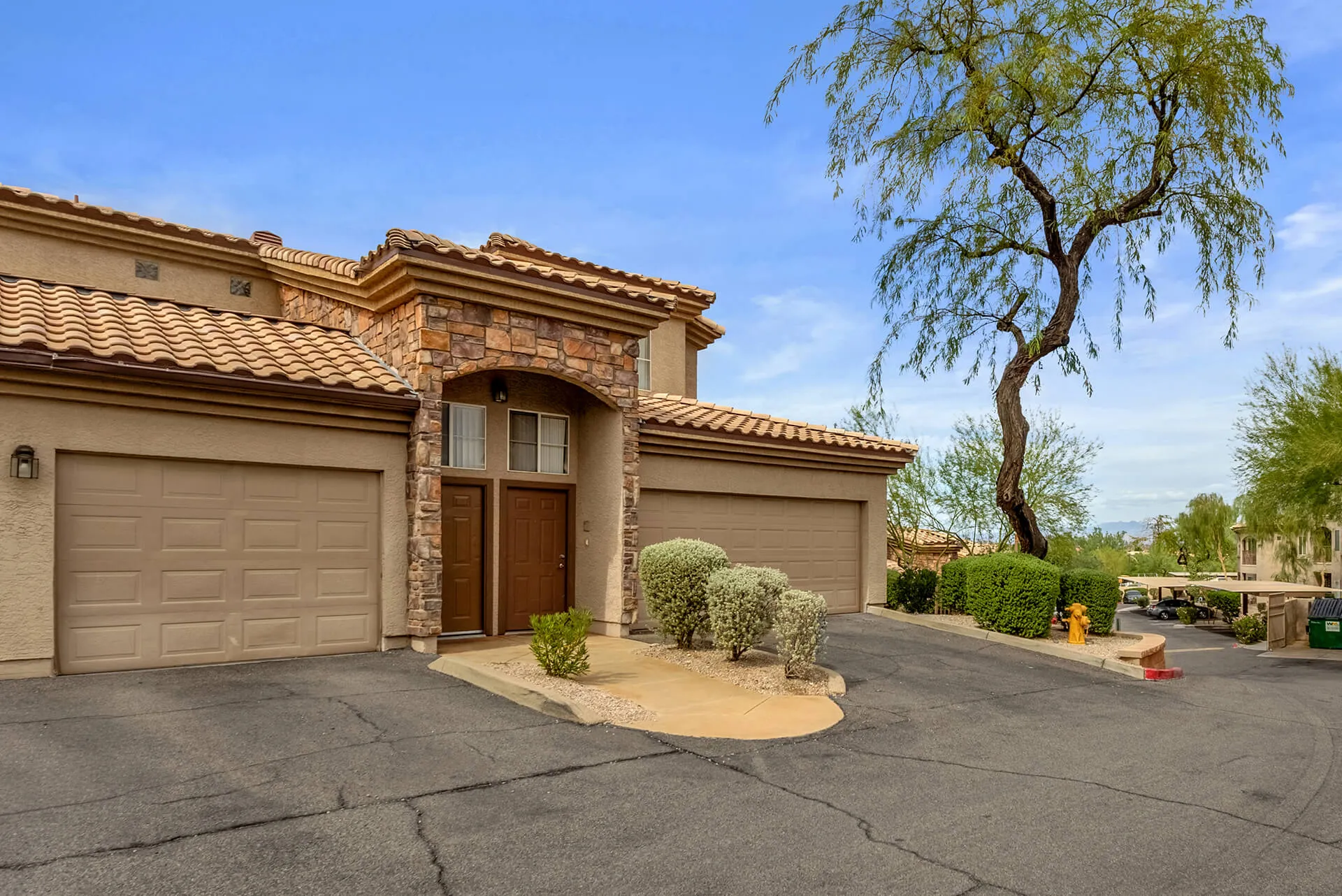 Modern two-story house with beige exterior, attached garage, landscaped front yard, large shade tree, and clear blue sky.
