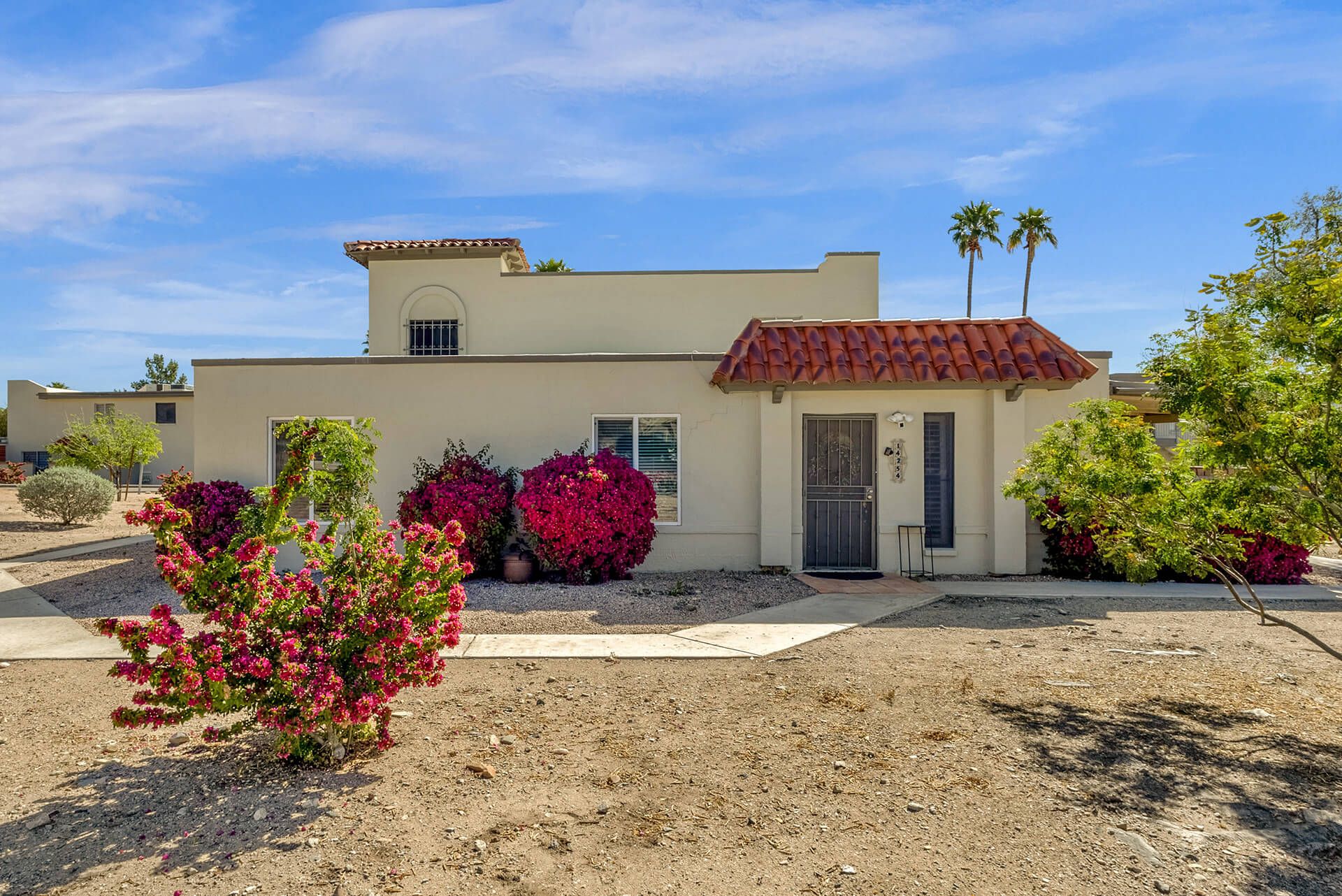 Modern two-story house with beige exterior, attached garage, landscaped front yard, large shade tree, and clear blue sky.