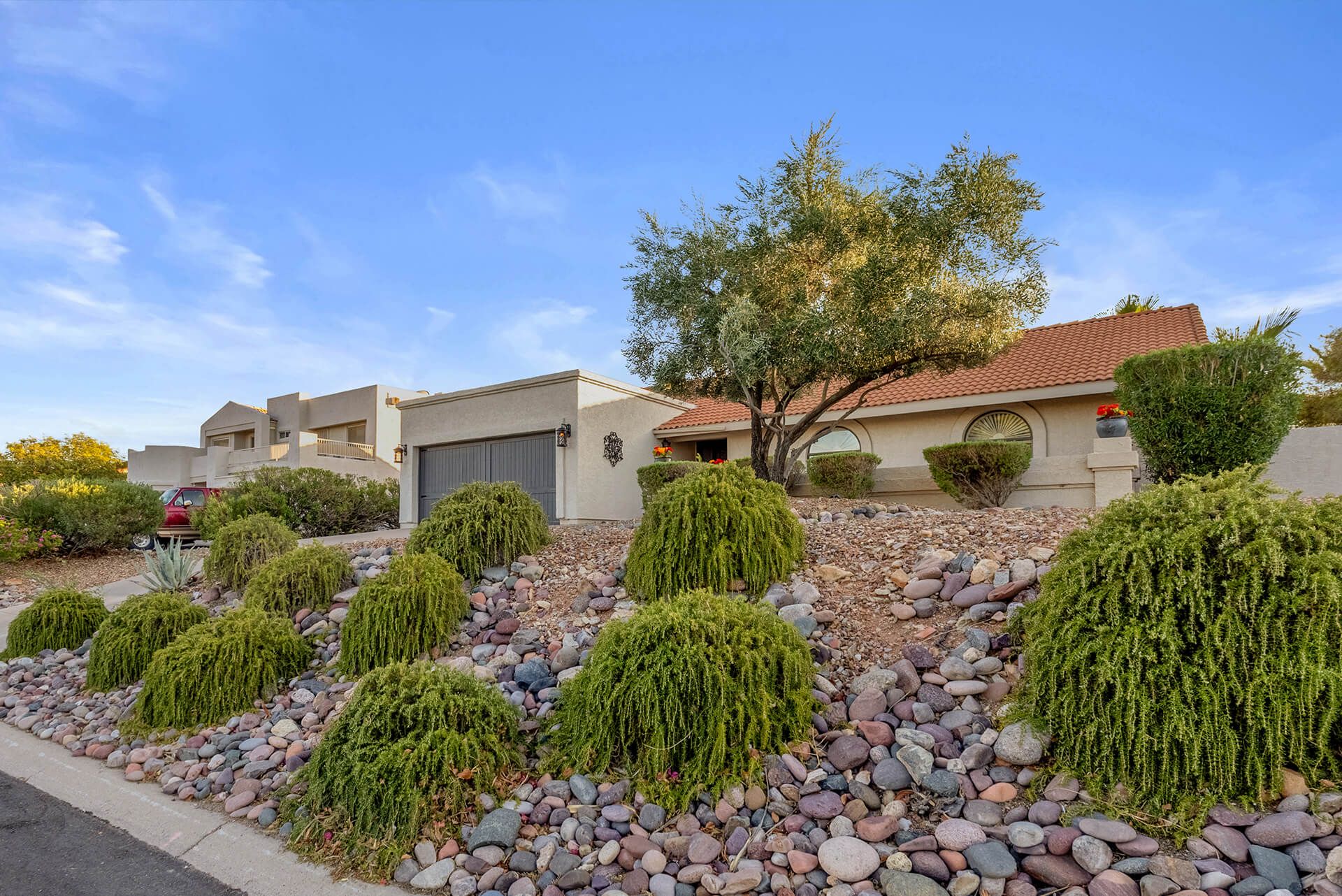 Single-story desert-style house with a tiled roof, surrounded by desert plants including cacti and a large tree, under a bright blue sky.