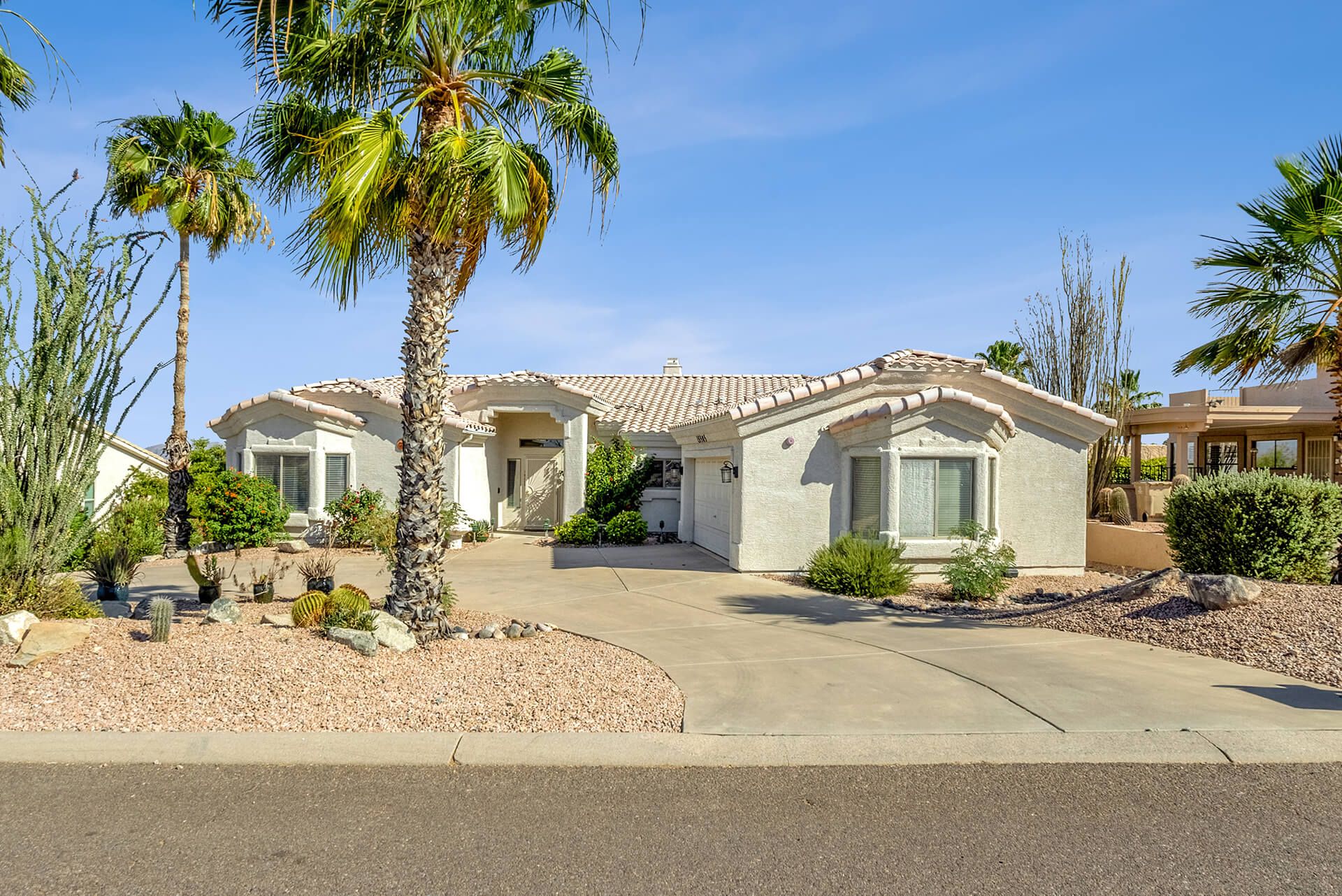Single-story desert-style house with a tiled roof, surrounded by desert plants including cacti and a large tree, under a bright blue sky.
