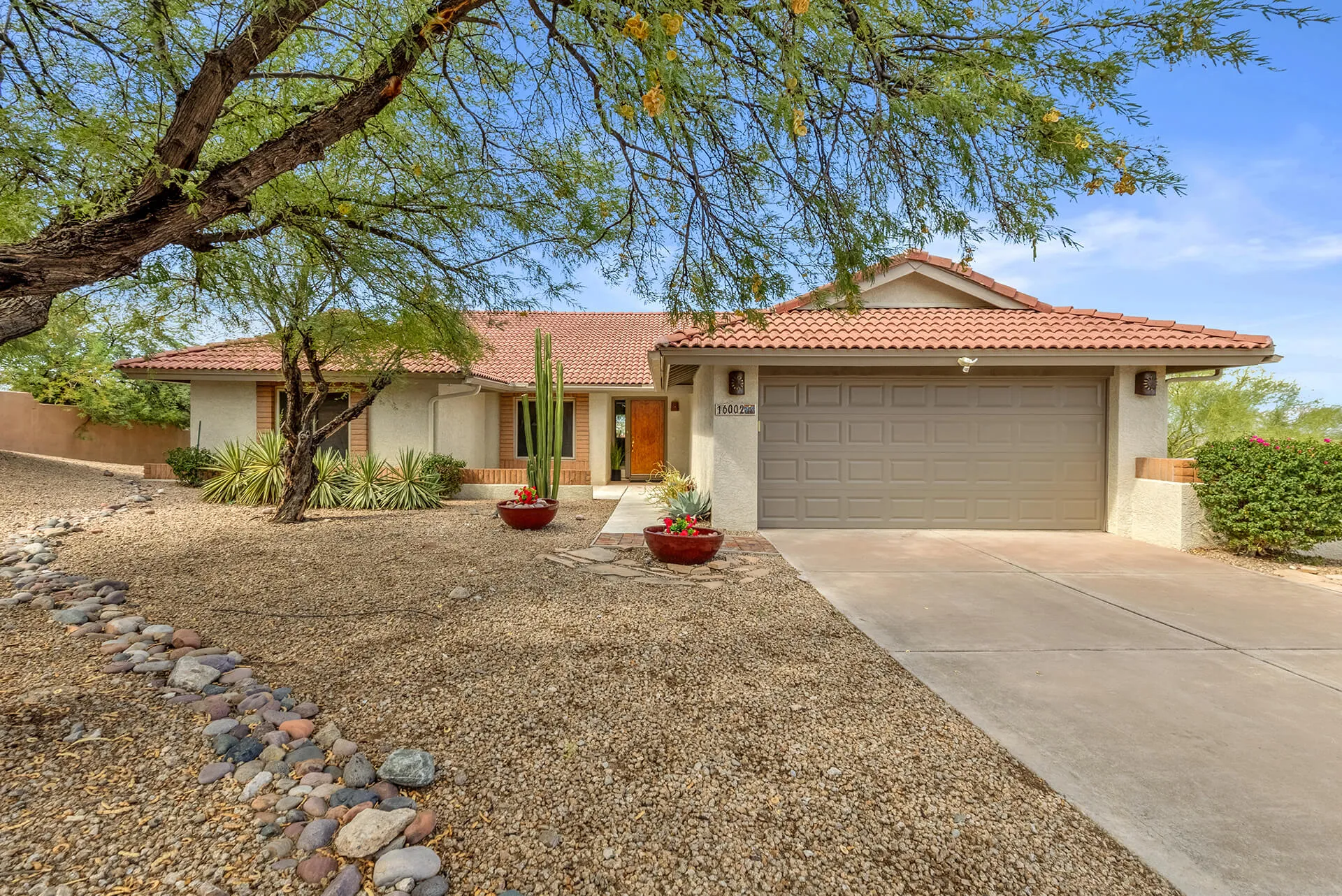 Single-story desert-style house with a tiled roof, surrounded by desert plants including cacti and a large tree, under a bright blue sky.