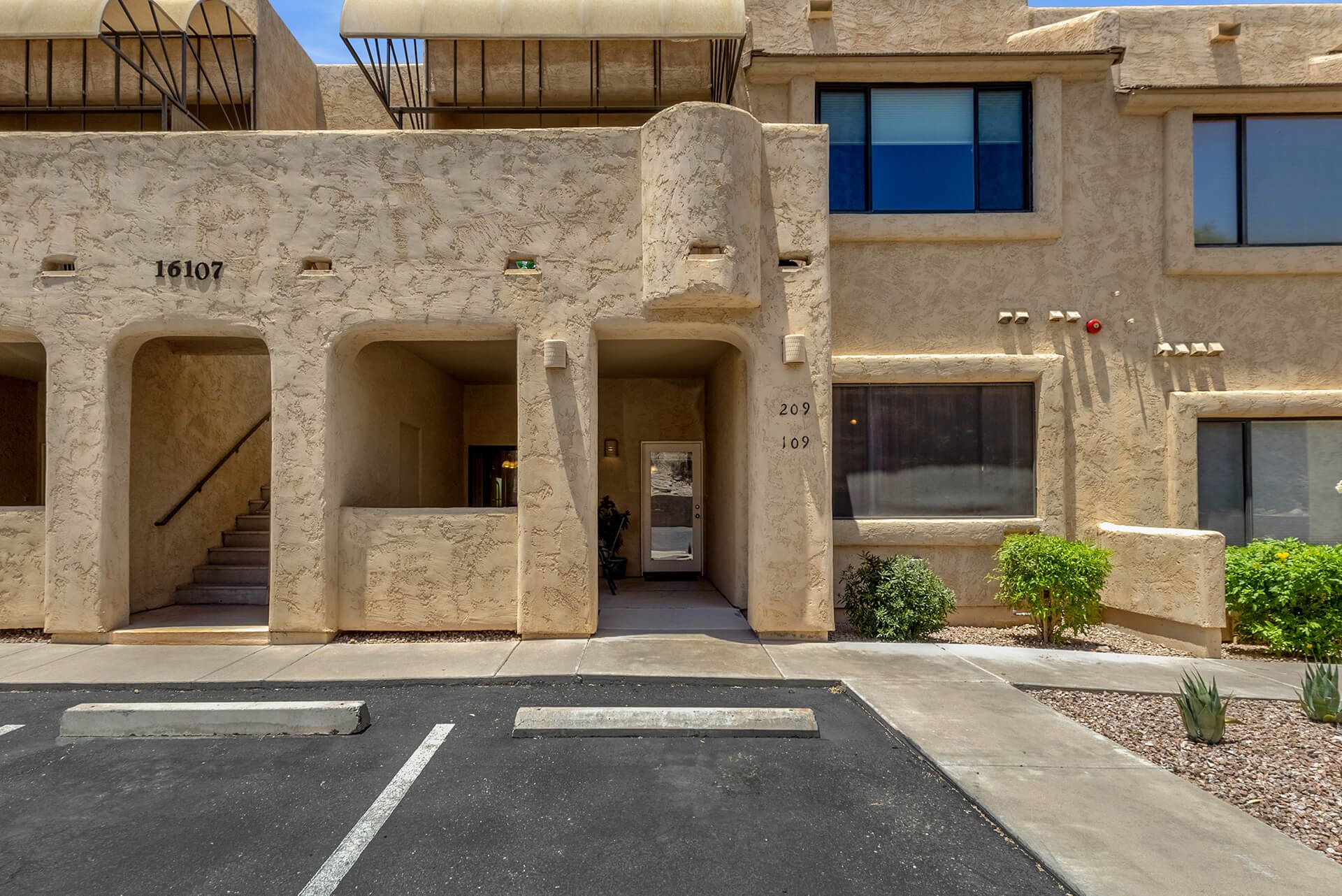Single-story desert-style house with a tiled roof, surrounded by desert plants including cacti and a large tree, under a bright blue sky.