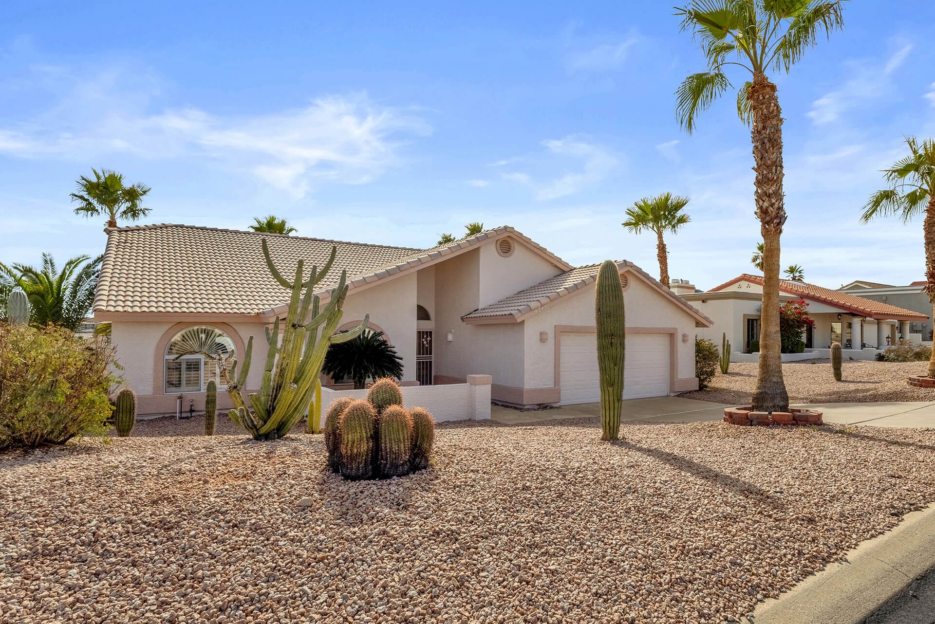 Modern two-story house with beige exterior, attached garage, landscaped front yard, large shade tree, and clear blue sky.