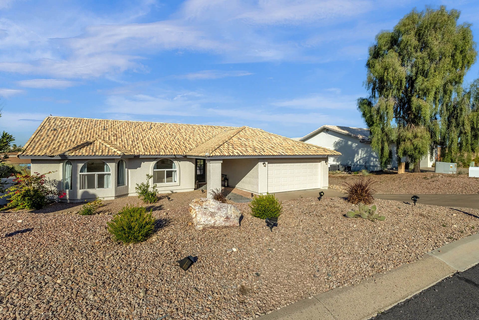 Modern two-story house with beige exterior, attached garage, landscaped front yard, large shade tree, and clear blue sky.