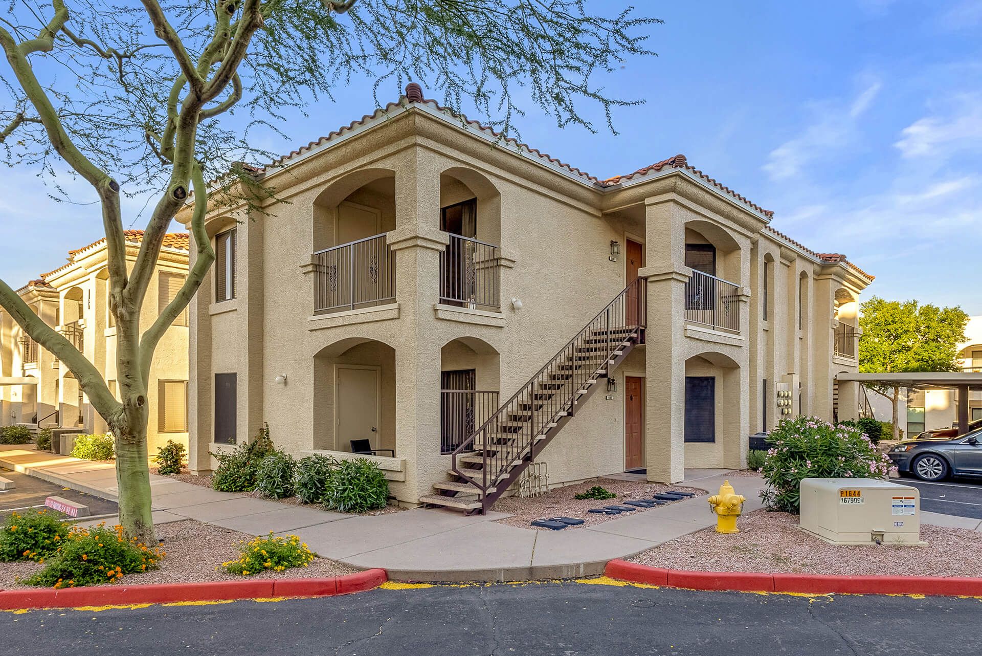 Modern two-story house with beige exterior, attached garage, landscaped front yard, large shade tree, and clear blue sky.