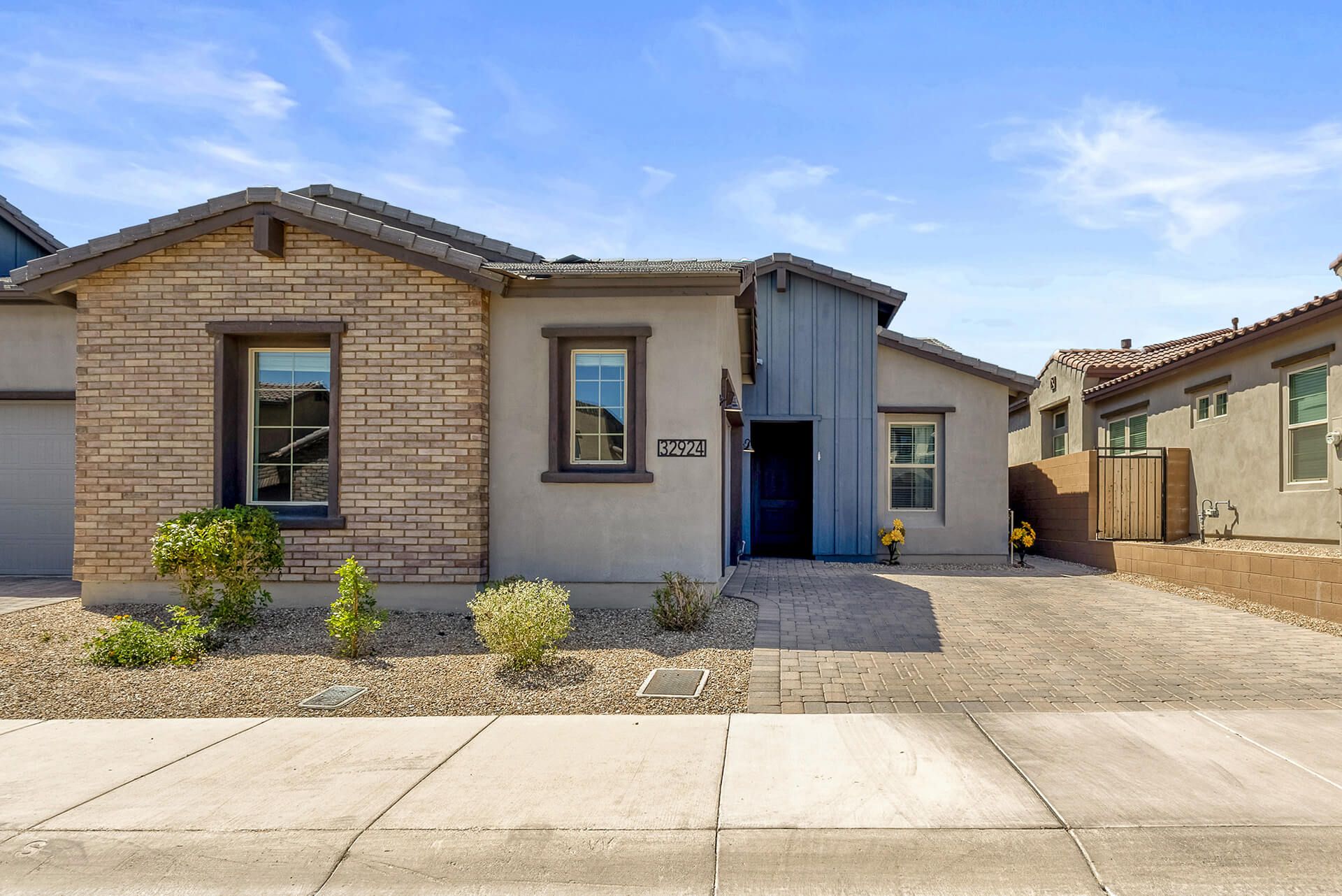 Modern two-story house with beige exterior, attached garage, landscaped front yard, large shade tree, and clear blue sky.