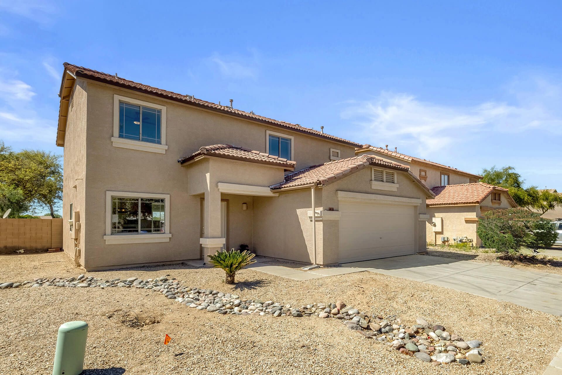 Modern two-story house with beige exterior, attached garage, landscaped front yard, large shade tree, and clear blue sky.