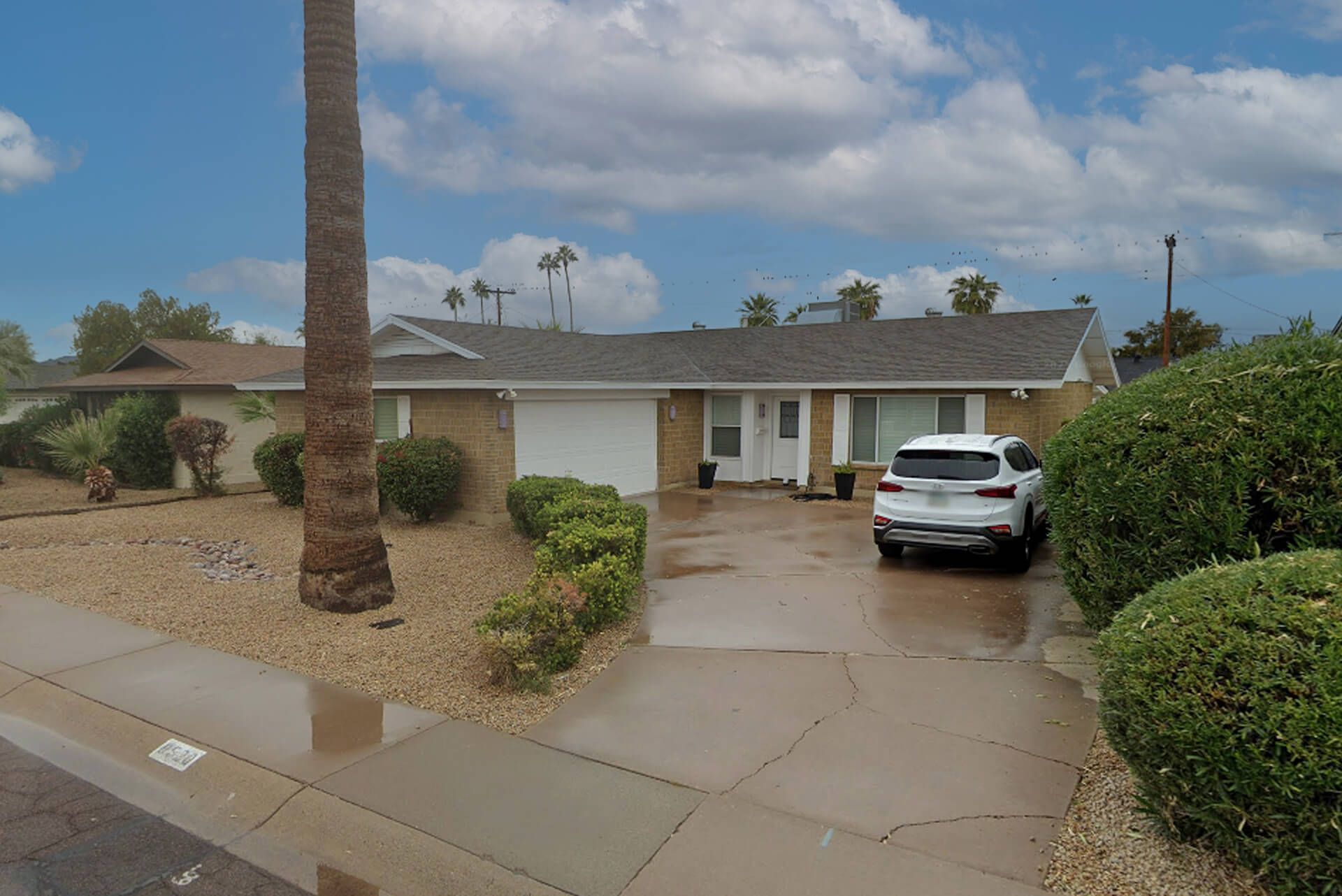 Single-story suburban house with a three-car garage, beige stucco exterior, tan tile roof, desert landscaping, large front windows, and a small landscaped area with rocks and shrubs, under a clear blue sky.