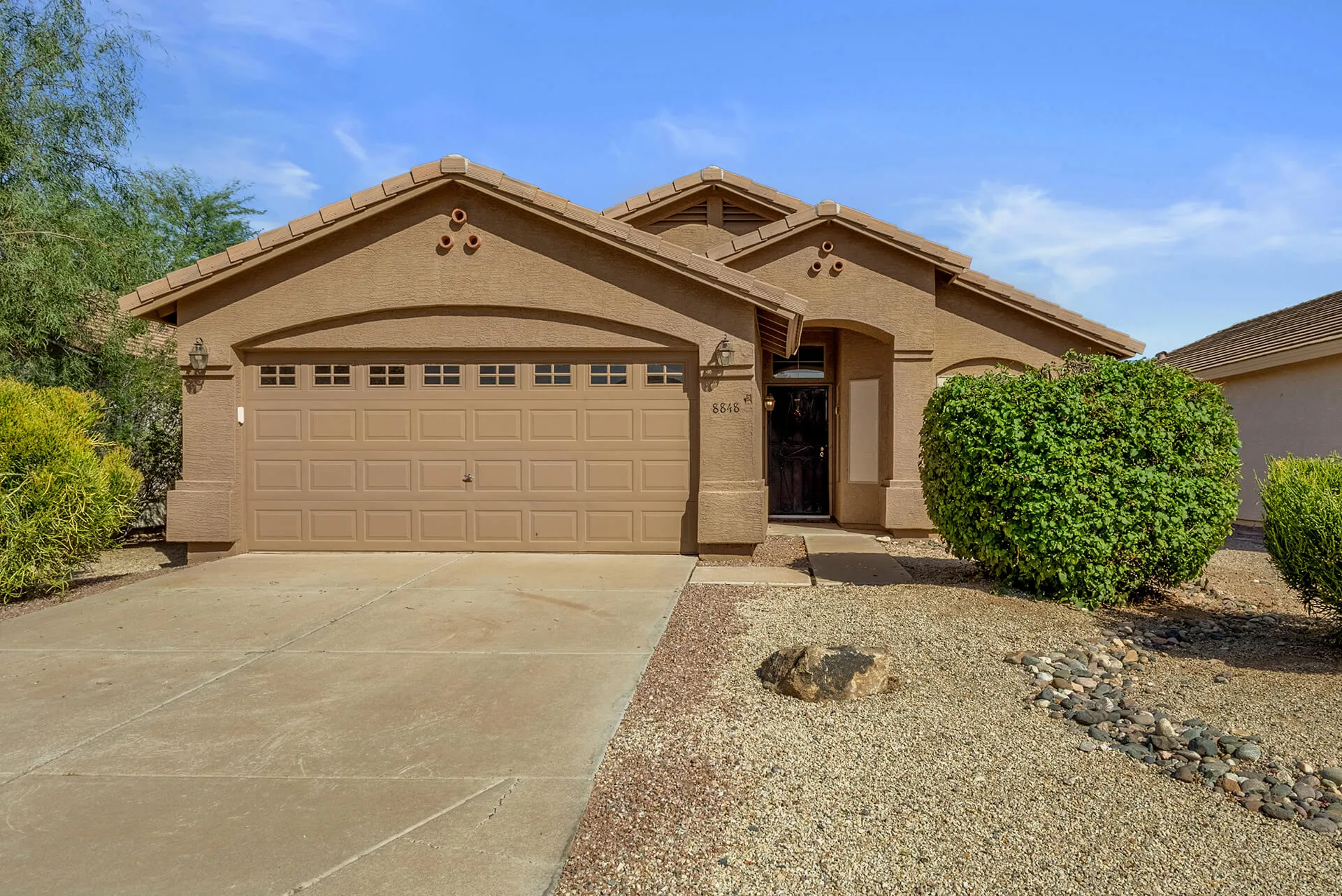 Modern two-story house with beige exterior, attached garage, landscaped front yard, large shade tree, and clear blue sky.