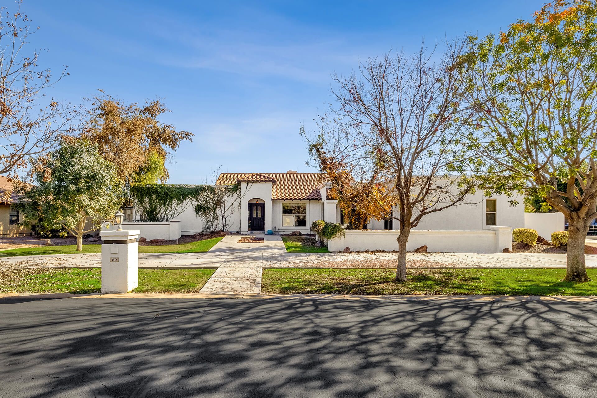 Single-story modern white house with a tiled roof, front yard with trees and green lawn, and clear blue sky in the background.