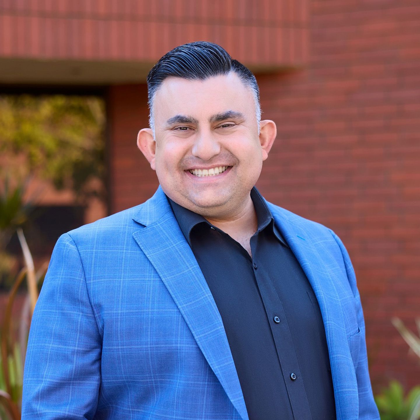Professional headshot of a smiling man in a black suit, white shirt, and orange tie against a plain white background
