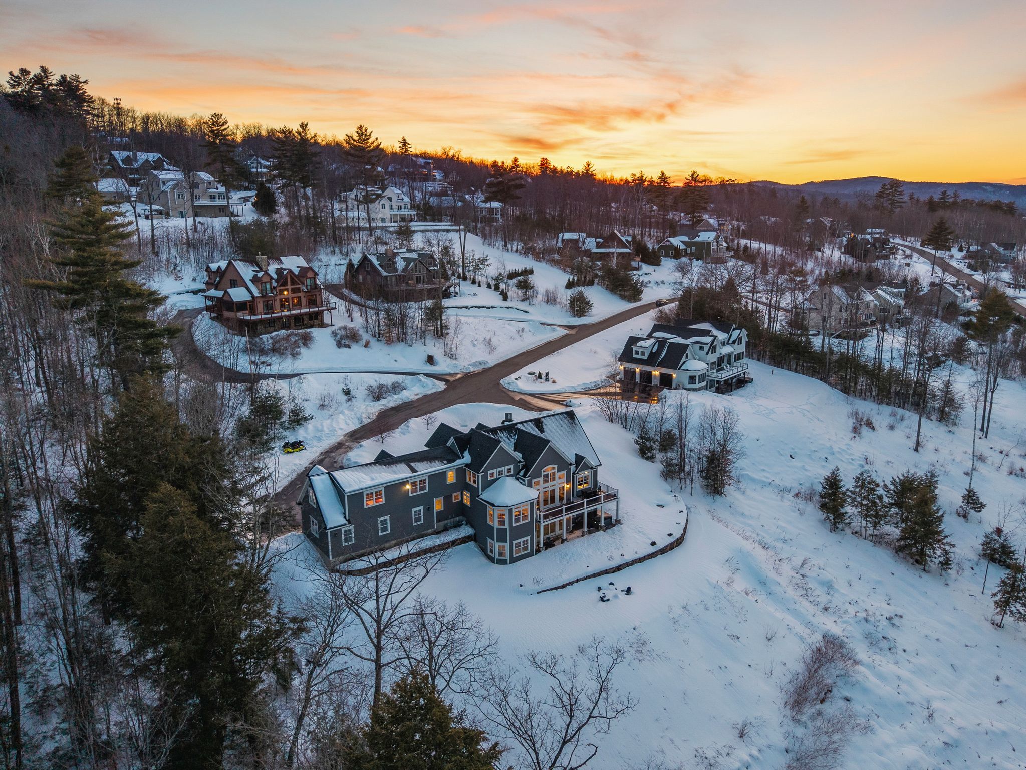 Spacious suburban house with beige siding, multiple windows, a garage door, surrounded by a lush green lawn, trees, and a scenic view of mountains and a river in the background on a partly cloudy day.