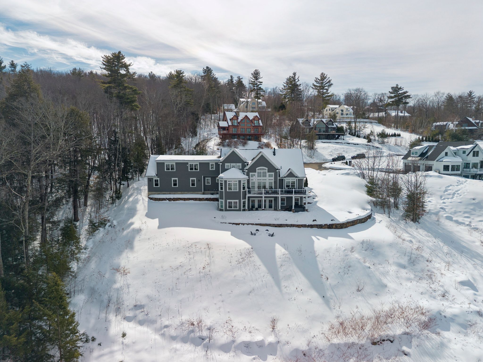Aerial view of a spacious house with a large deck, illuminated at dusk, nestled among lush green trees in a scenic suburban neighborhood with other houses and a winding road.