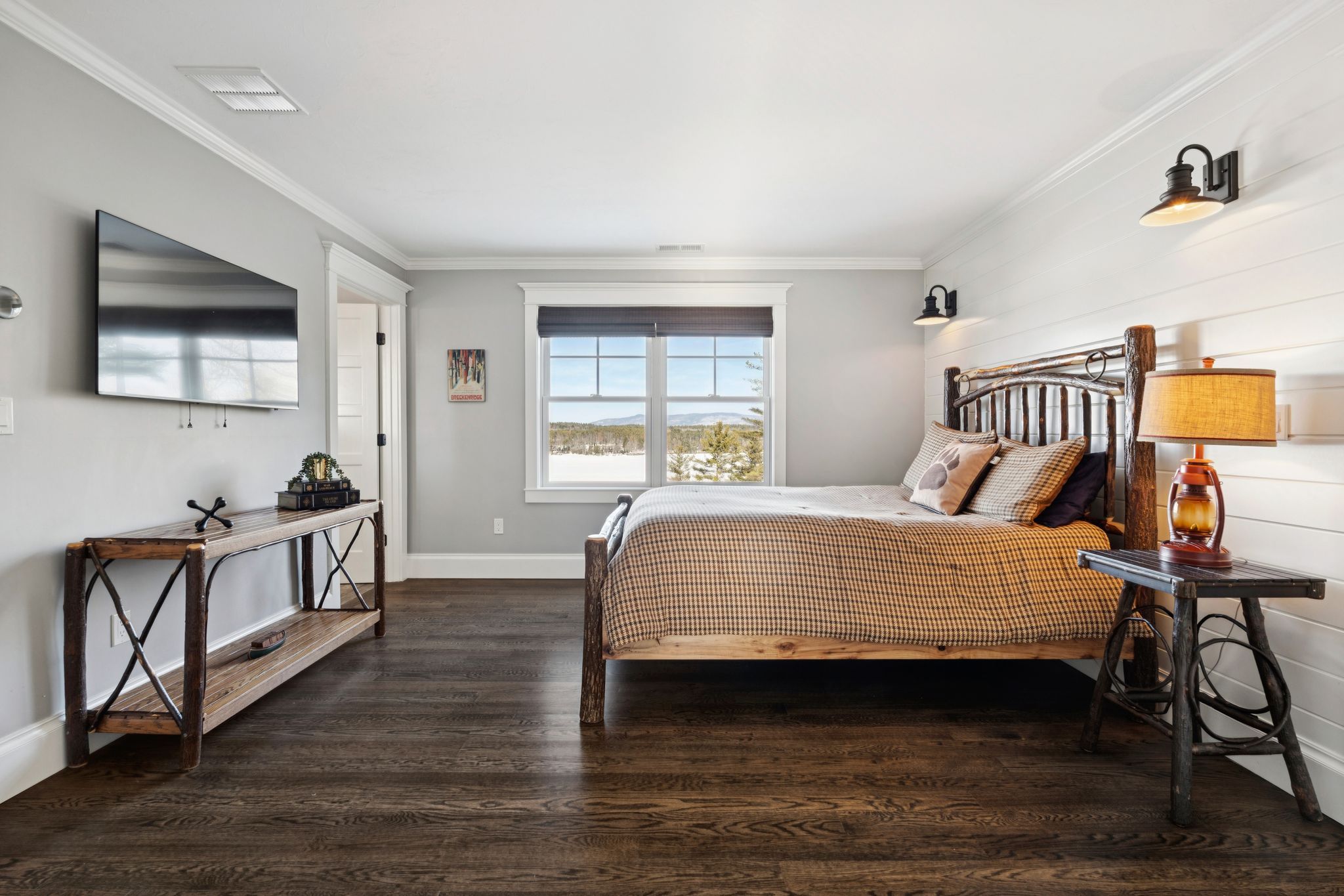 Cozy home office with a daybed, plaid bedding, front and window-side desks with monitors, white cabinets, and a patterned rug in a room with green walls and large windows overlooking a lush outdoor area.