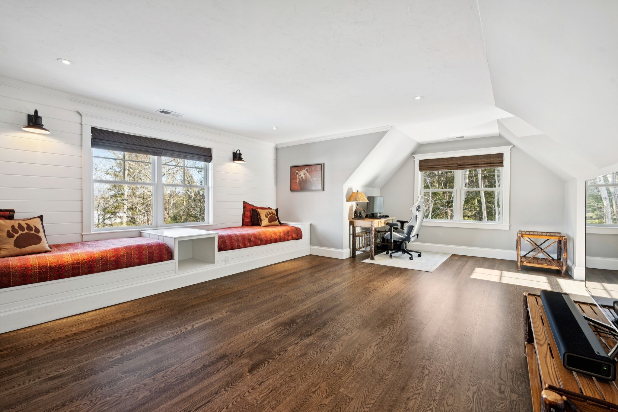 Cozy bedroom with king-sized bed, dark gray bedding, white pillows, wooden nightstands with lamps, wall-mounted mountain artwork, window with lush green trees outside, and rustic wooden ski patrol sign.