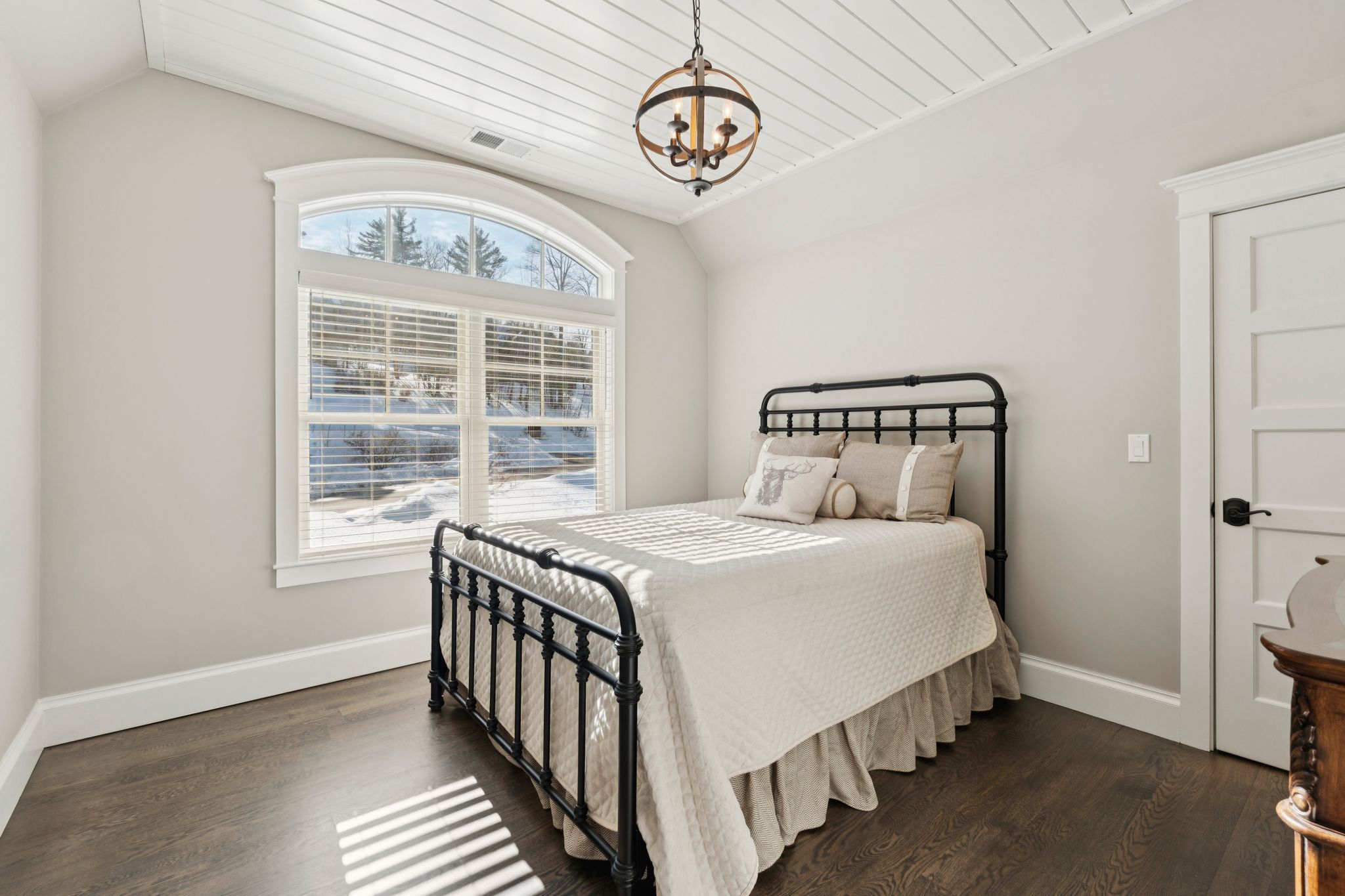 Cozy bedroom with wooden bunk beds, large arched window with white curtains, neutral wall color, ceiling fan, and decorative wall shelves with artwork and plants.