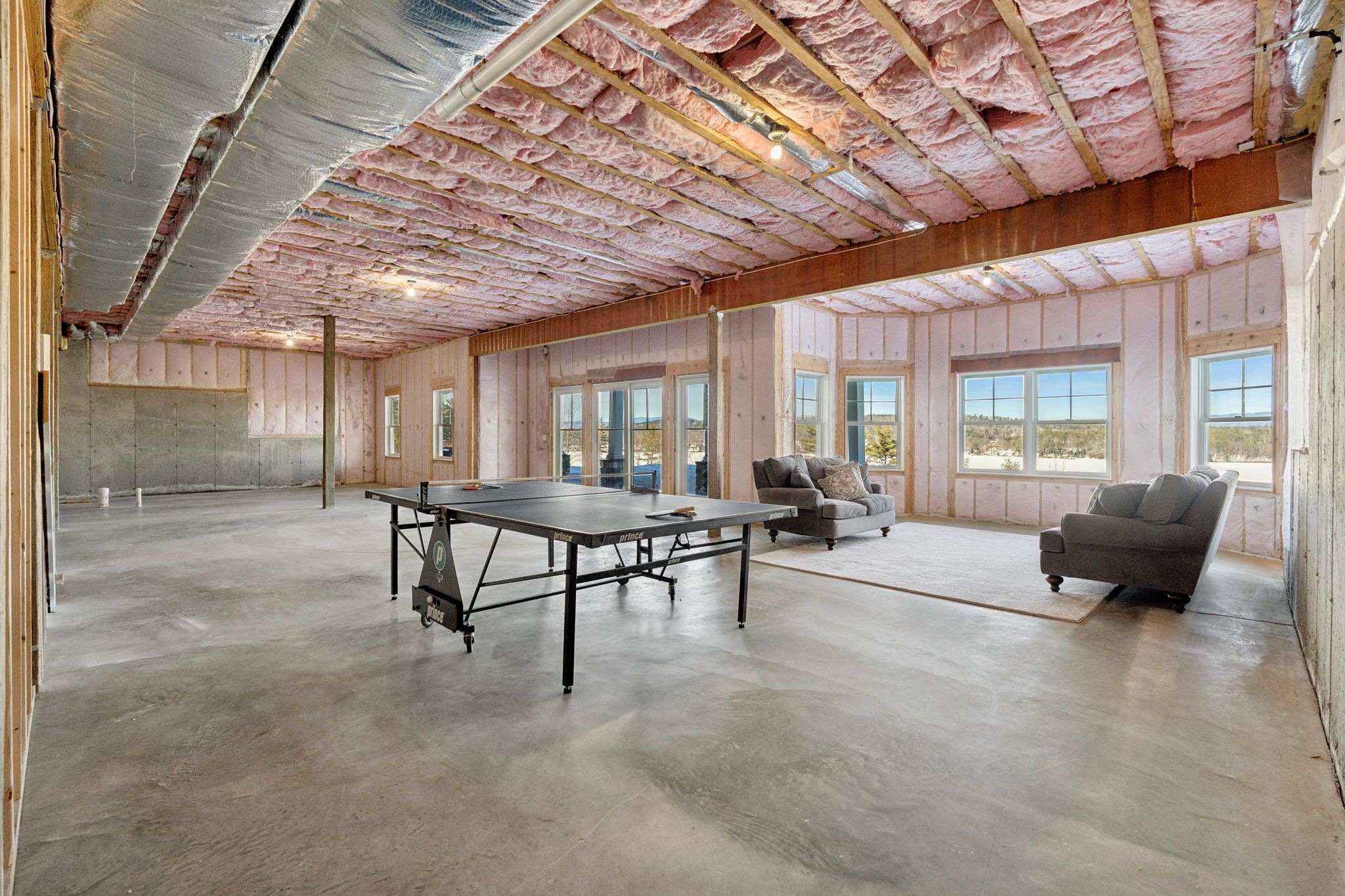 Spacious basement game room with shuffleboard, pool table, and seating area, featuring large windows and hardwood floors.