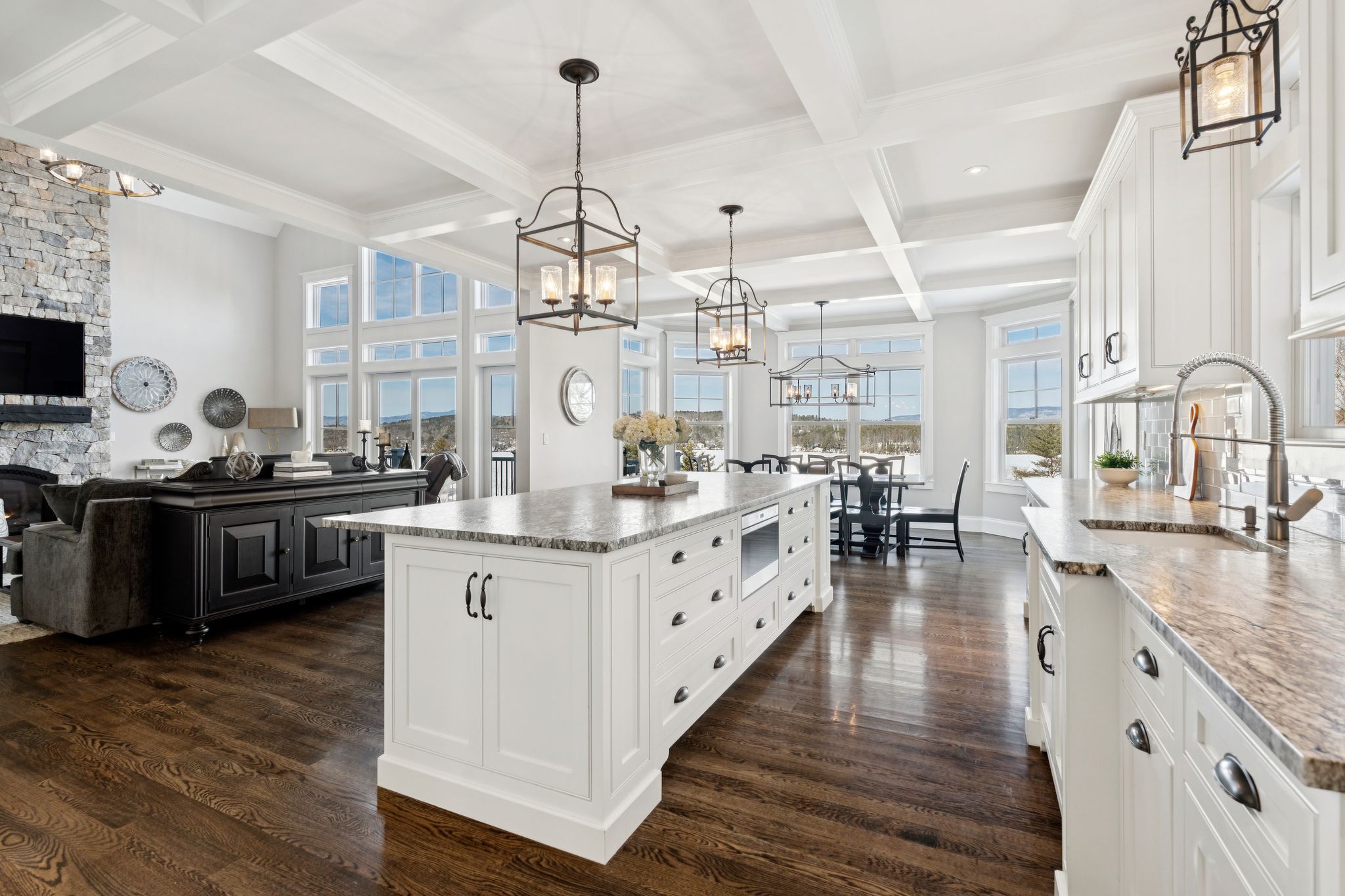 Modern kitchen with blue island and barstools, wooden cabinetry, mosaic backsplash, and large windows letting in natural light, decorated with flowers and seaside artwork.