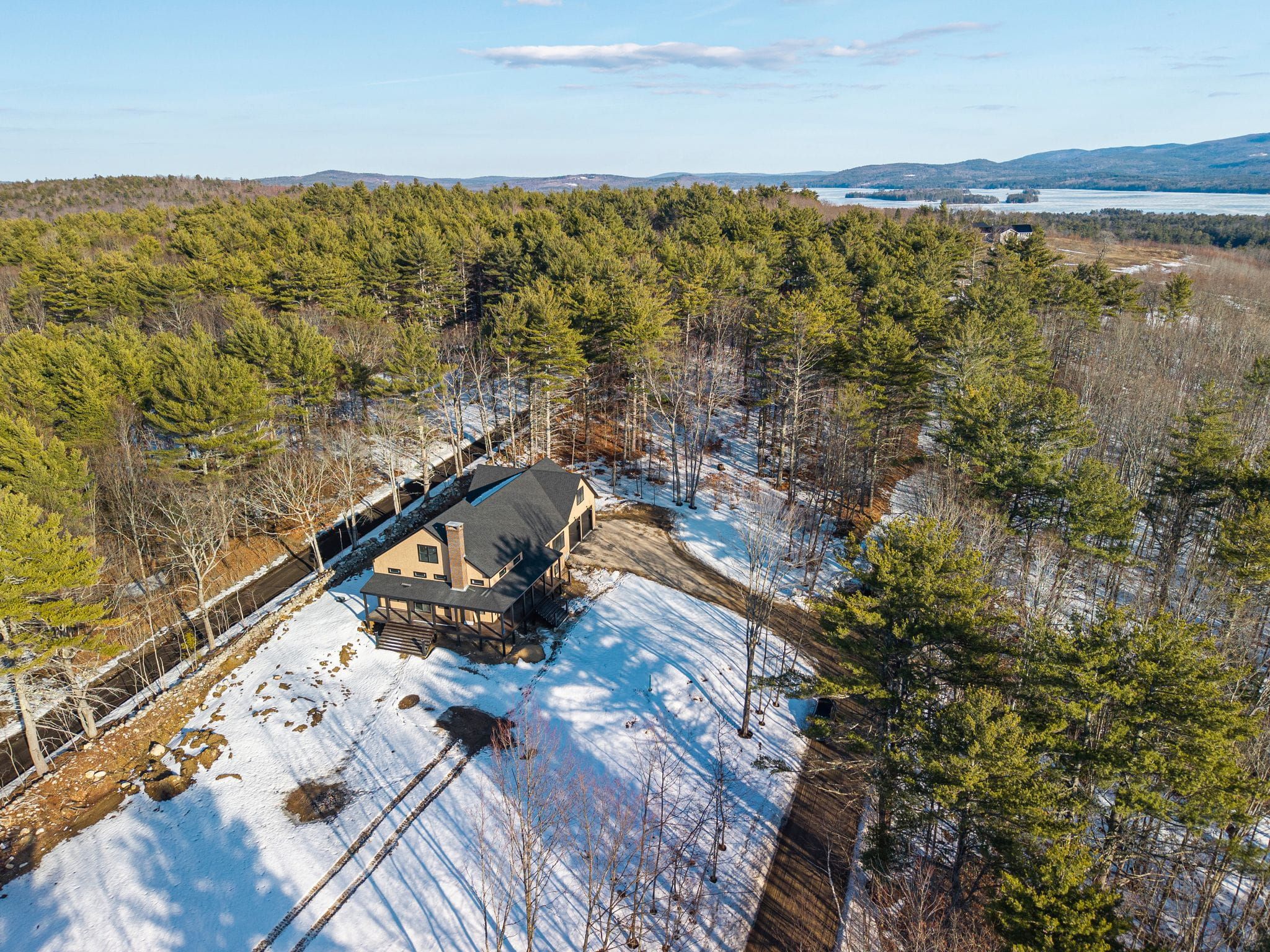 Snow-covered lakeside house with large windows, wooden decks, and surrounding trees in winter scene