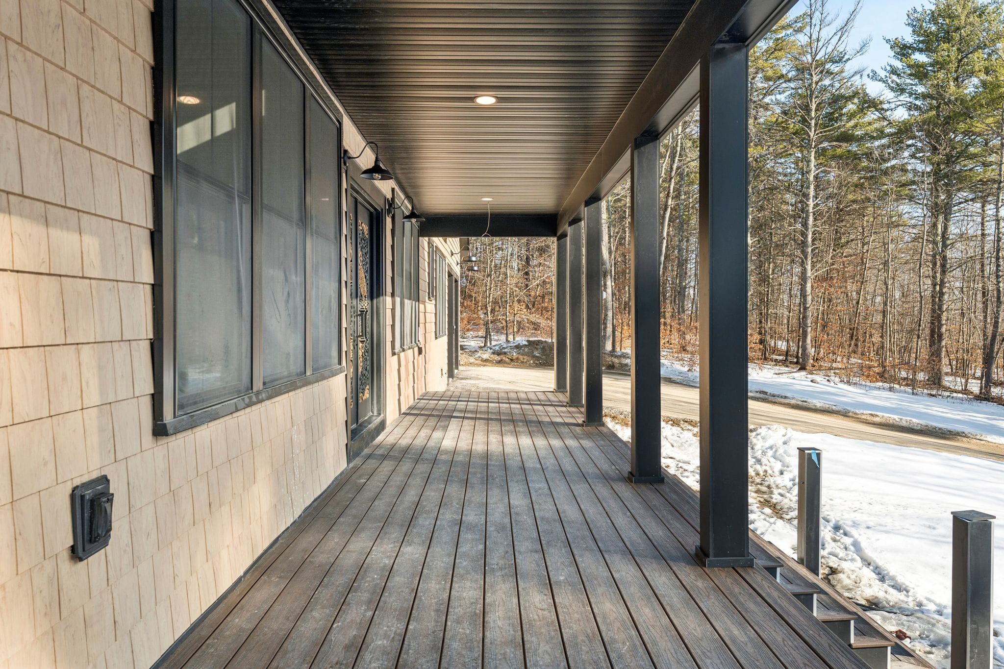 Modern lakeside house with large windows and illuminated interior, surrounded by snow-covered landscape and tall pine trees during winter evening.