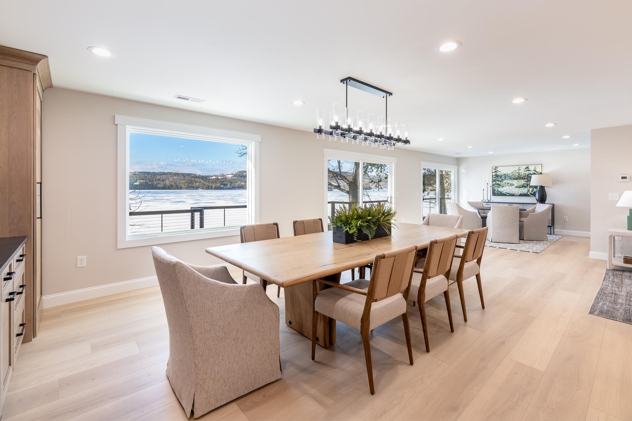 Lakeside house with a wooden dock and kayaks on a sandy beach, surrounded by tall pine trees during sunset.