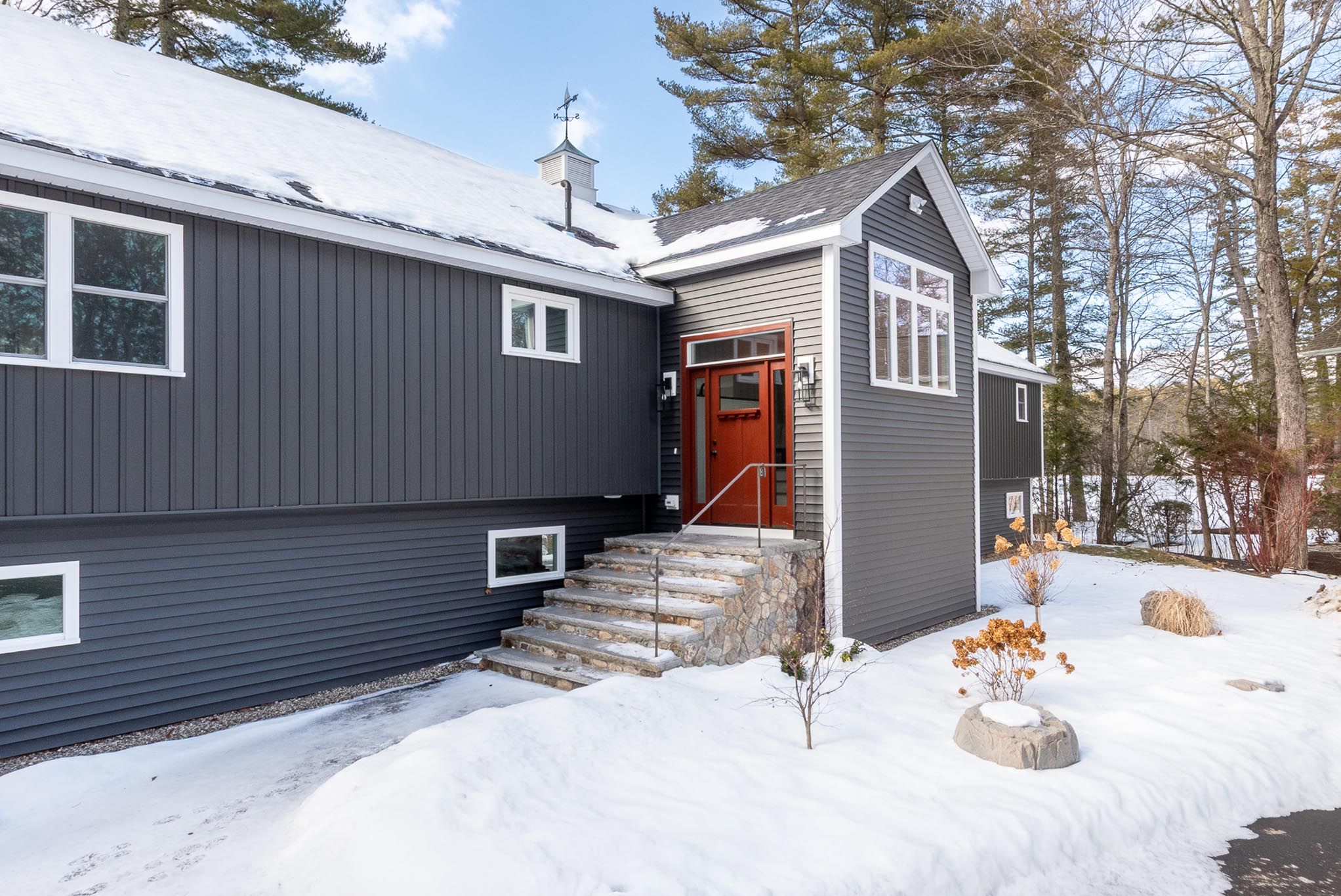 Modern lakeside house with a large deck, kayak and paddleboards on sandy beach, surrounded by lush green trees.