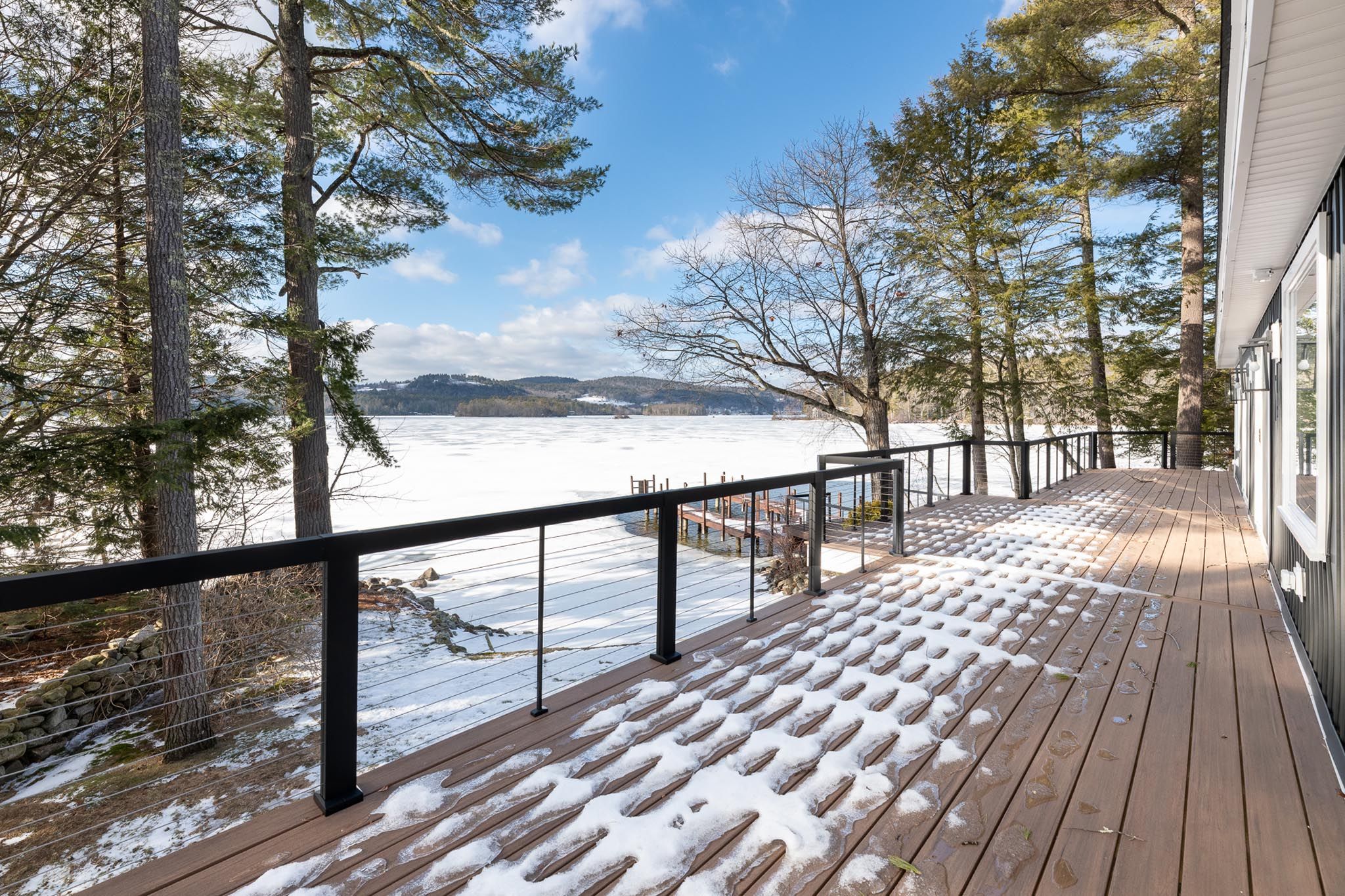 Modern lakeside house with a wooden dock and kayaks, surrounded by lush green trees and clear water.