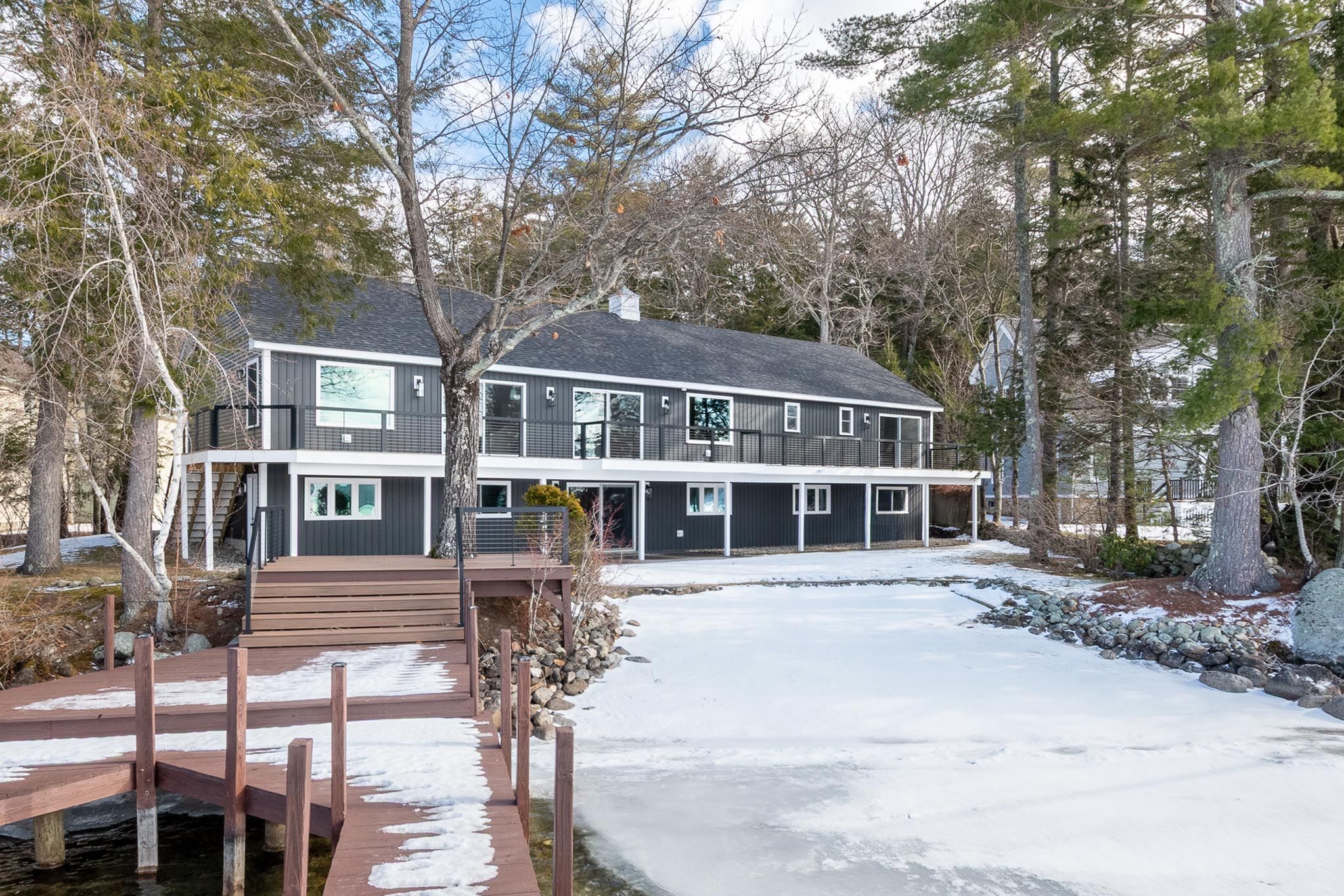 Modern dark gray two-story house with a well-maintained front yard, surrounded by tall trees and a clear blue sky with sunlight shining through.