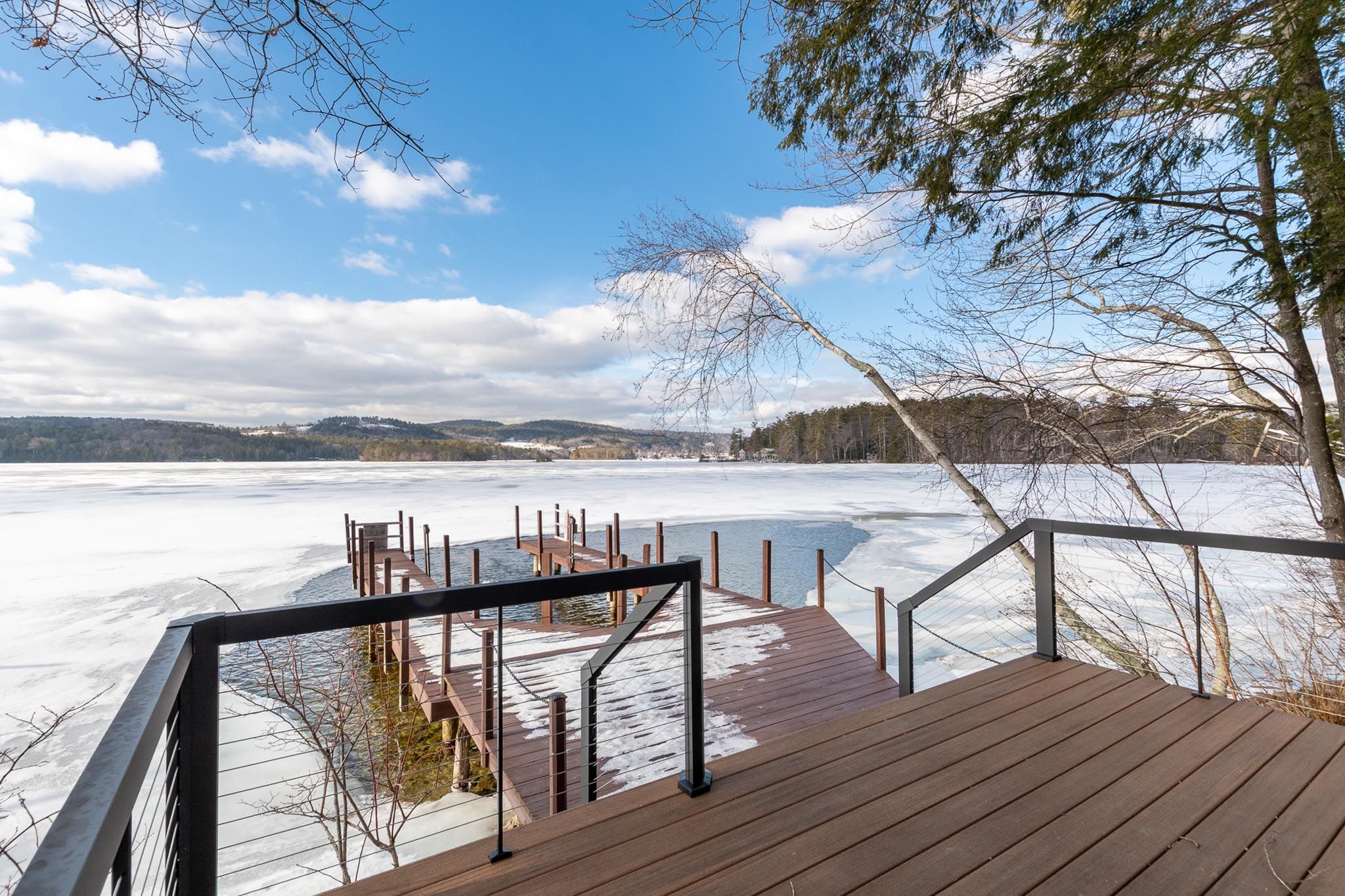 Modern wooden deck overlooking a lake with lush green trees and clear blue sky, with sunlight filtering through the leaves