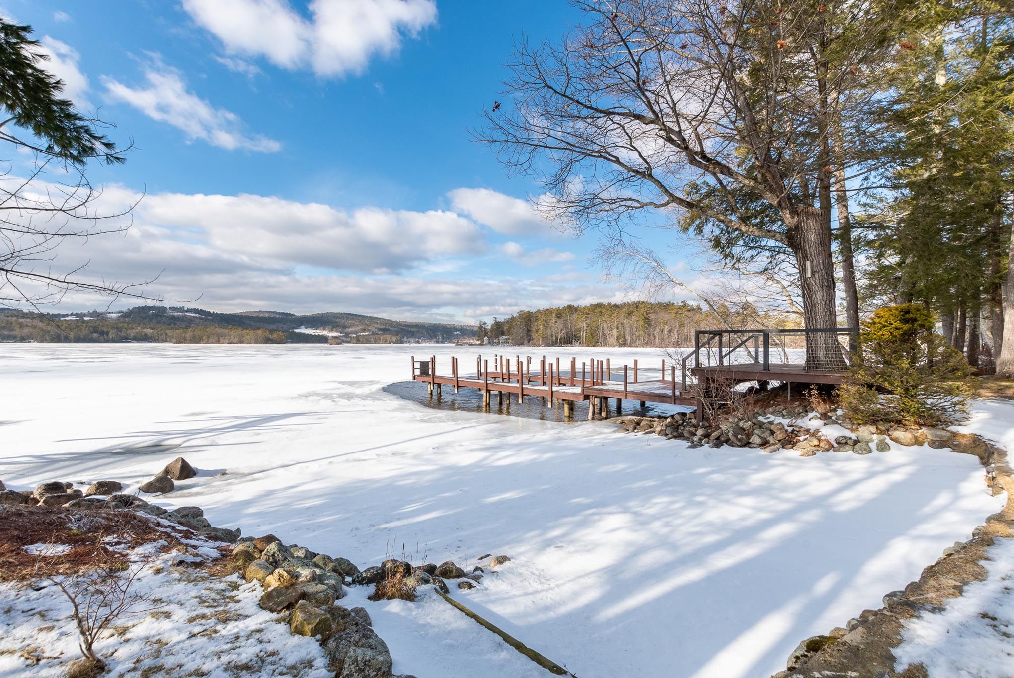 Beautiful lakeside deck with black metal railing, surrounded by lush green trees and views of the water, featuring sliding glass doors and outdoor wall-mounted lights for relaxing outdoor living.