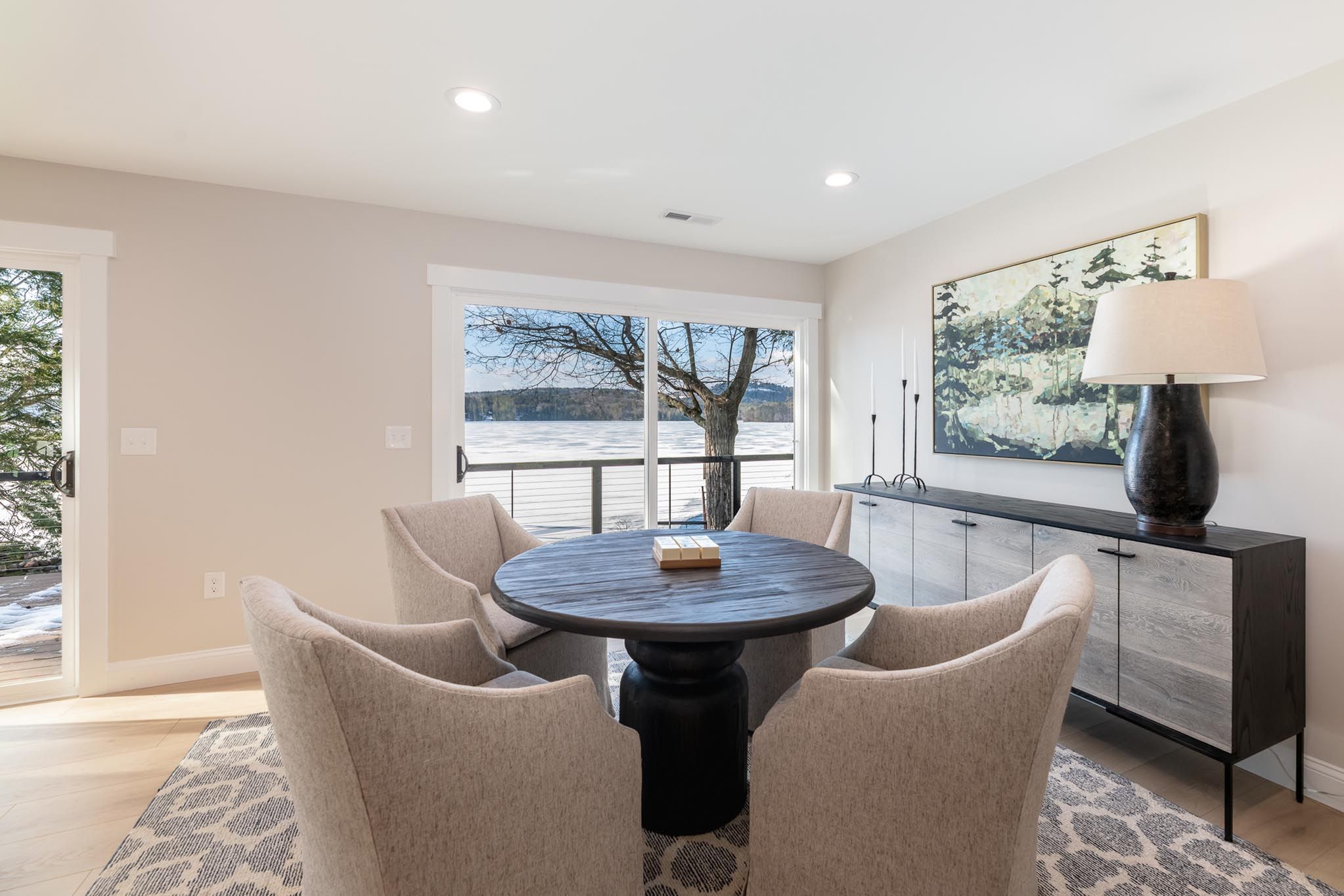 Aerial view of a lakeside house surrounded by tall green trees with a dock extending into the water, boats anchored near the shoreline, and a colorful sunset sky in the background.