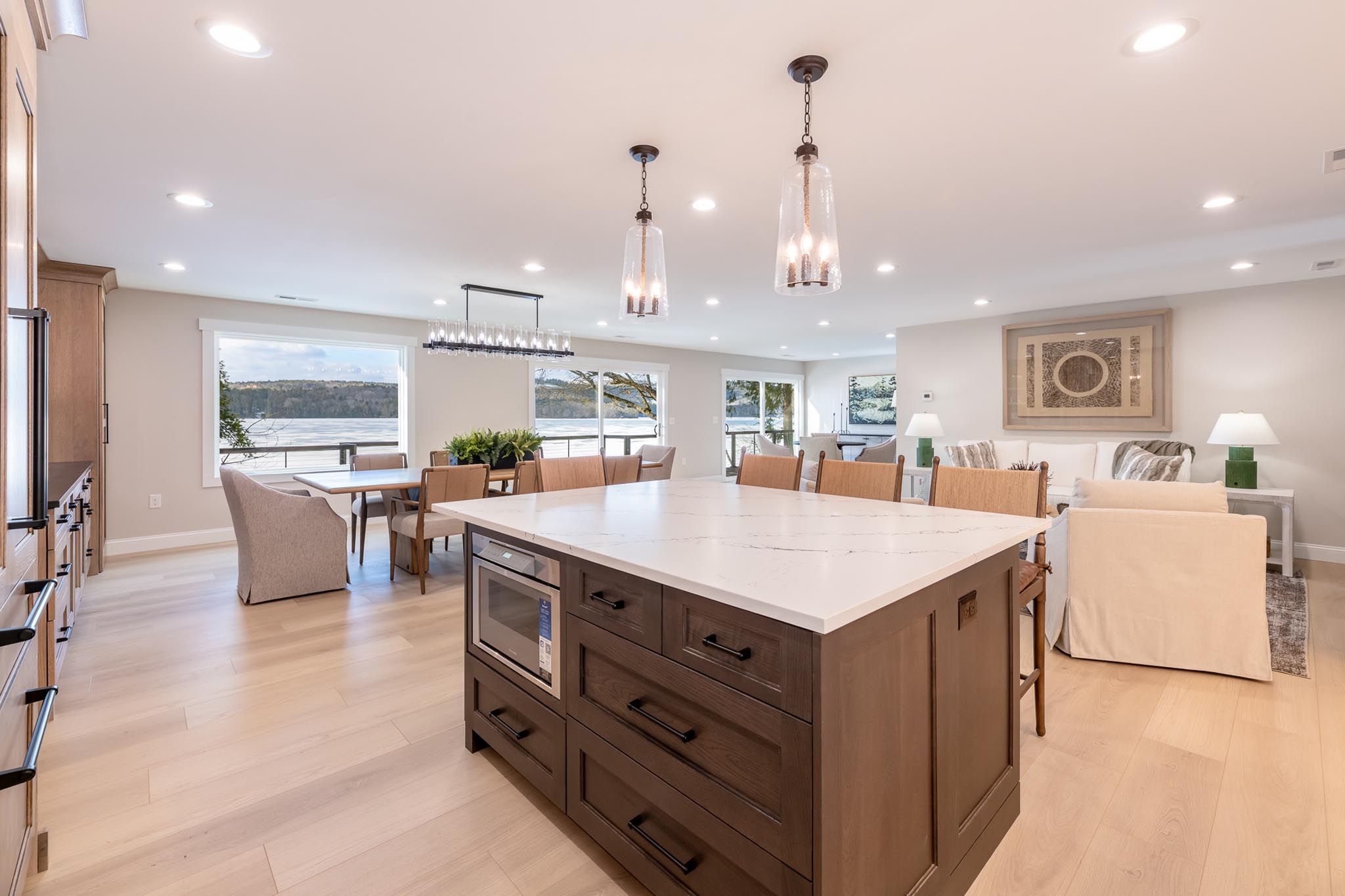 Modern two-story lakeside house with large glass windows, outdoor deck, lush greenery, kayaks on shore, and illuminated interior at dusk.