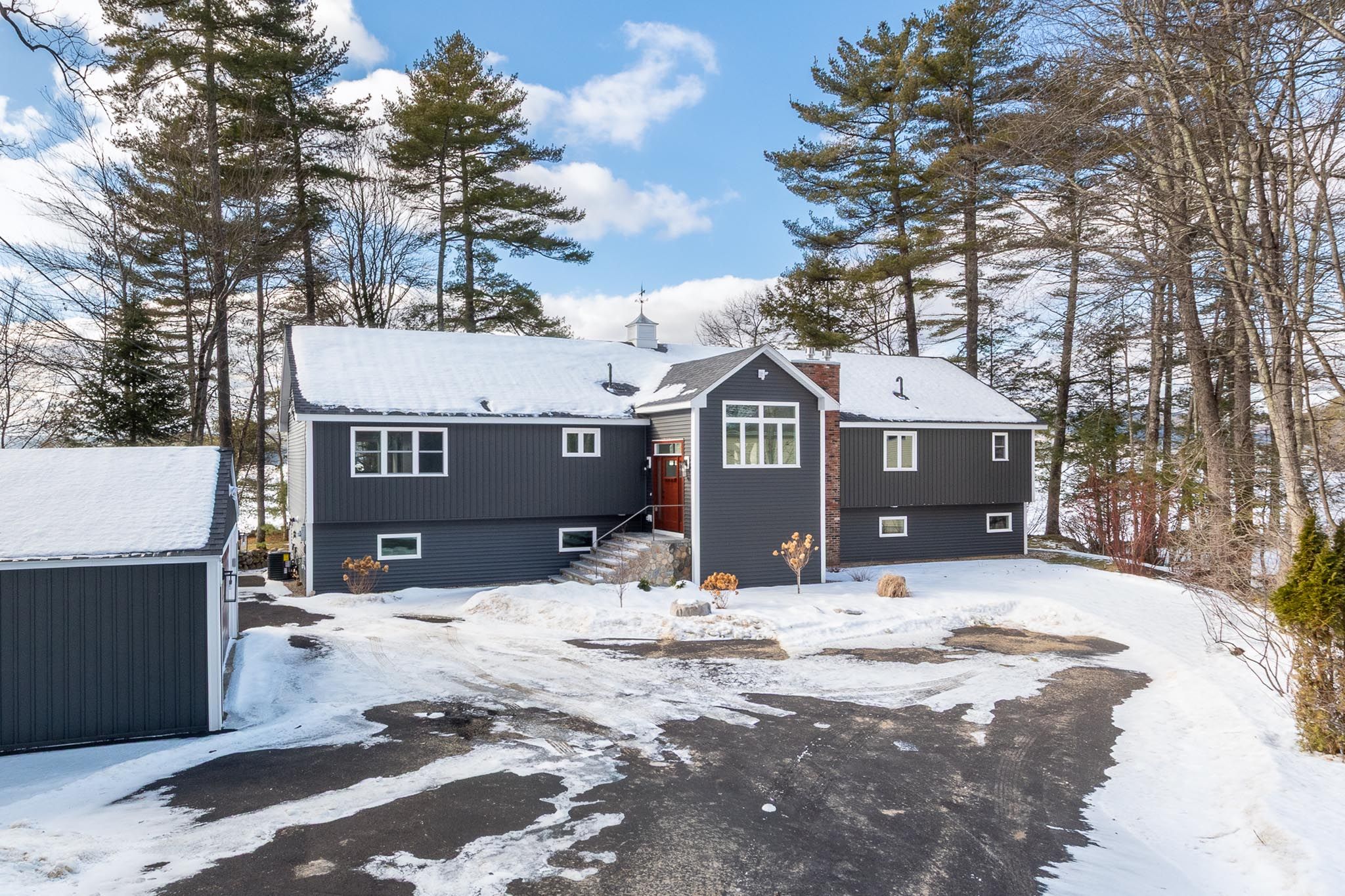 Lakeside house with a wooden dock, surrounded by lush green trees and water, featuring a black exterior with a balcony, situated in a natural wooded setting.