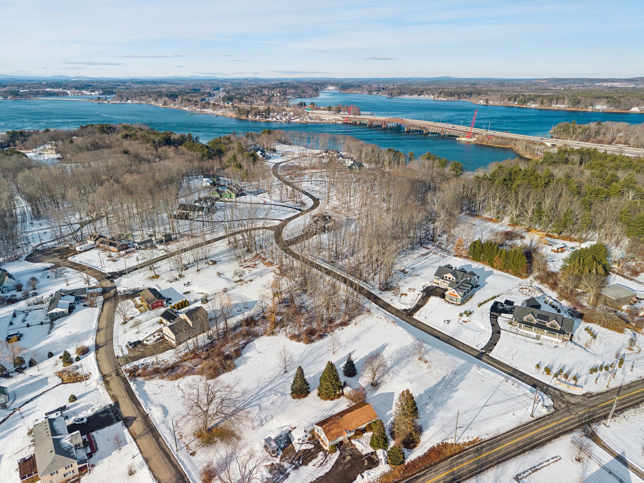 Aerial view of a snow-covered residential neighborhood near a large blue lake with a bridge crossing over the water, surrounded by trees and mountains in the distance.