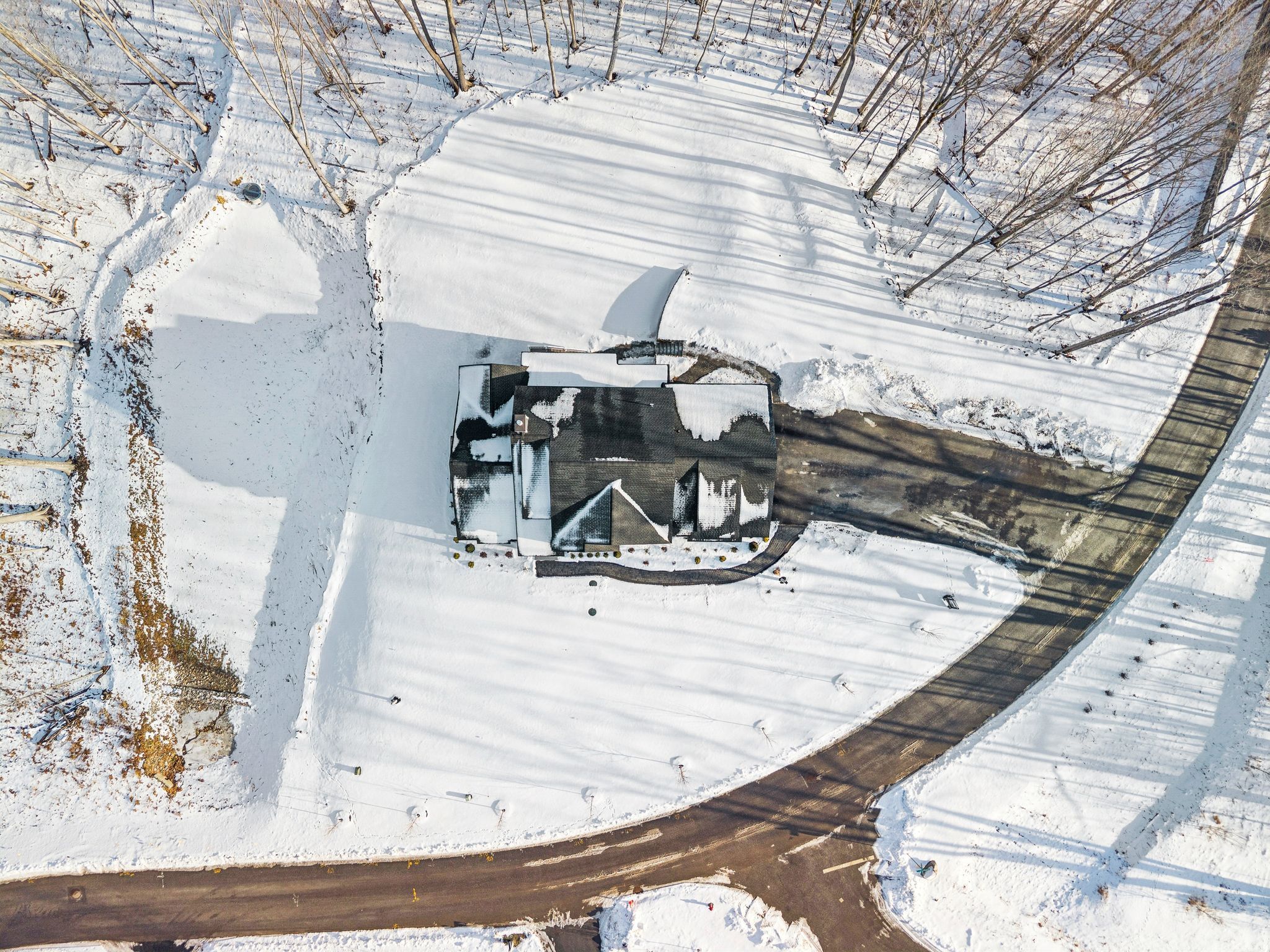 Aerial view of a snow-covered house surrounded by leafless trees and a winding driveway on a winter day