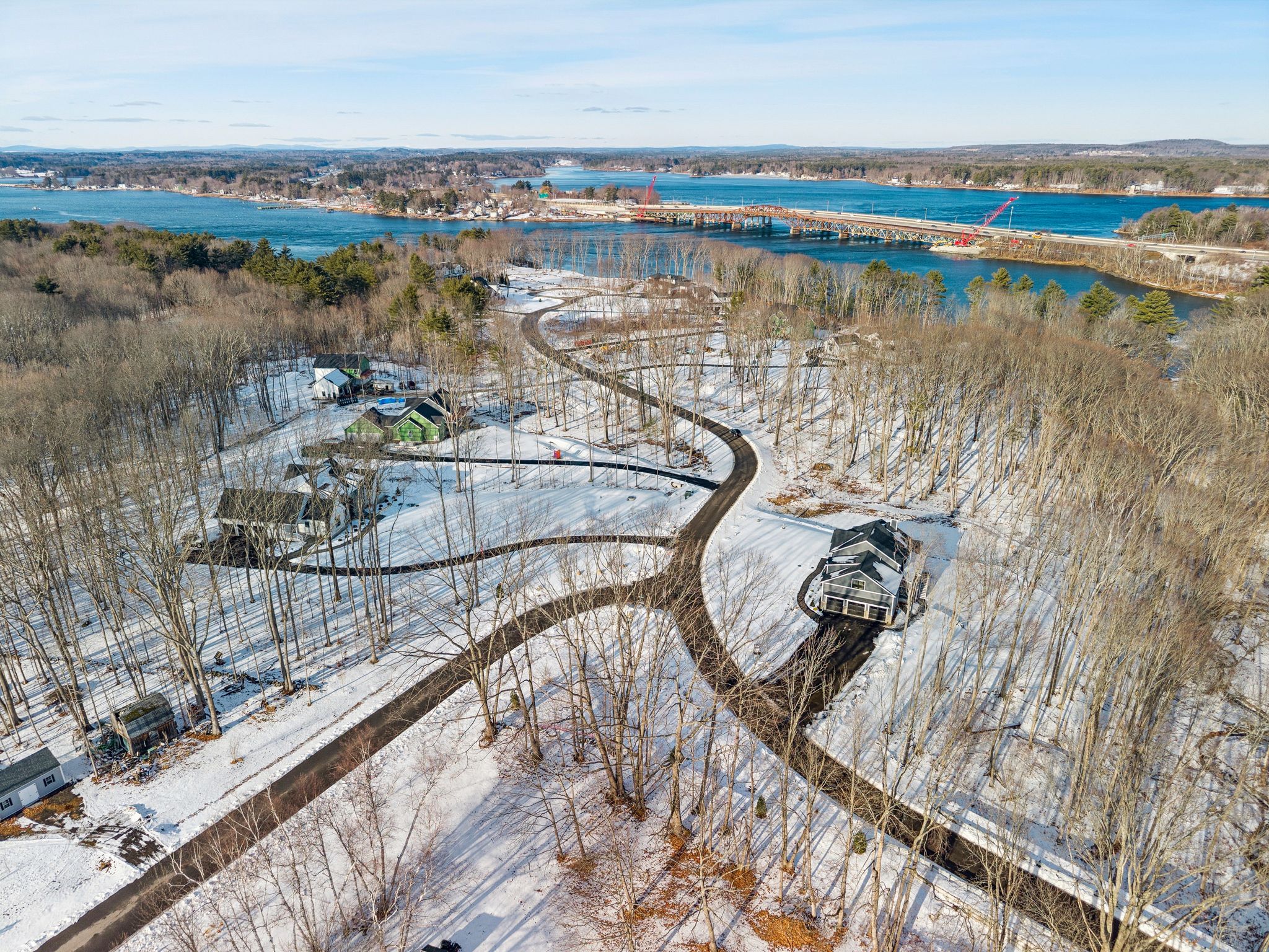Aerial view of a snowy residential area near a river with a bridge in the background and a winding driveway leading to houses surrounded by leafless trees.